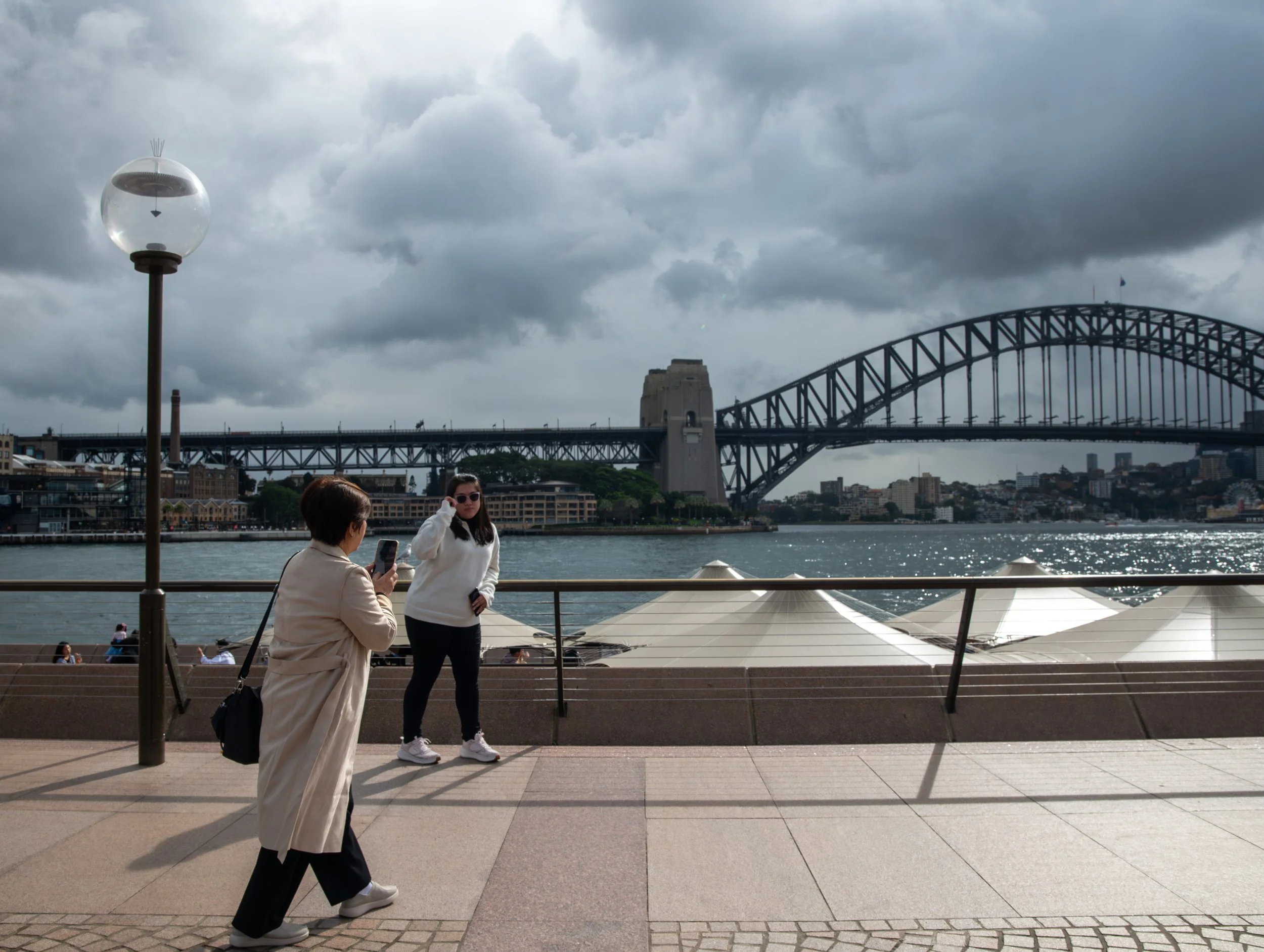 Two women near a waterfront with the Sydney Harbour Bridge in the background. One woman is taking a photo with her phone, the other is talking on a cell phone. Overcast skies with dark clouds.