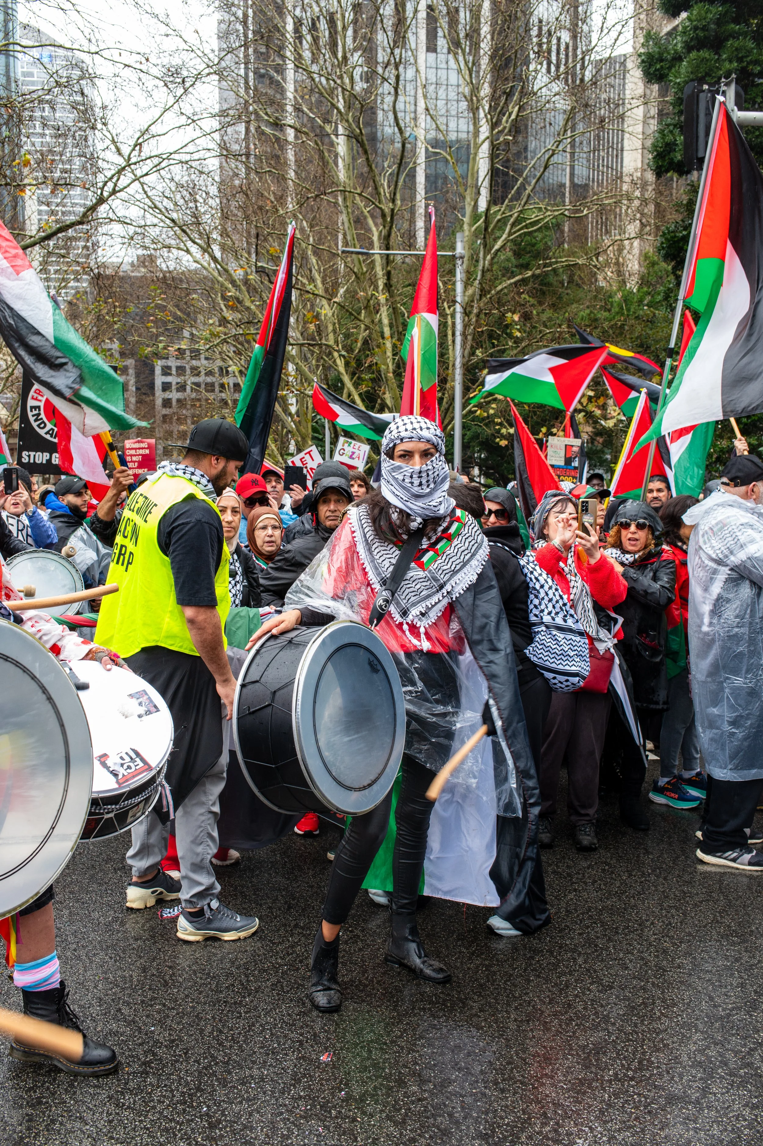 Protesters gathered in a city street holding Palestinian flags, with some playing drums, wearing headscarves, raincoats, and a yellow vest, amidst tall buildings and leafless trees.
