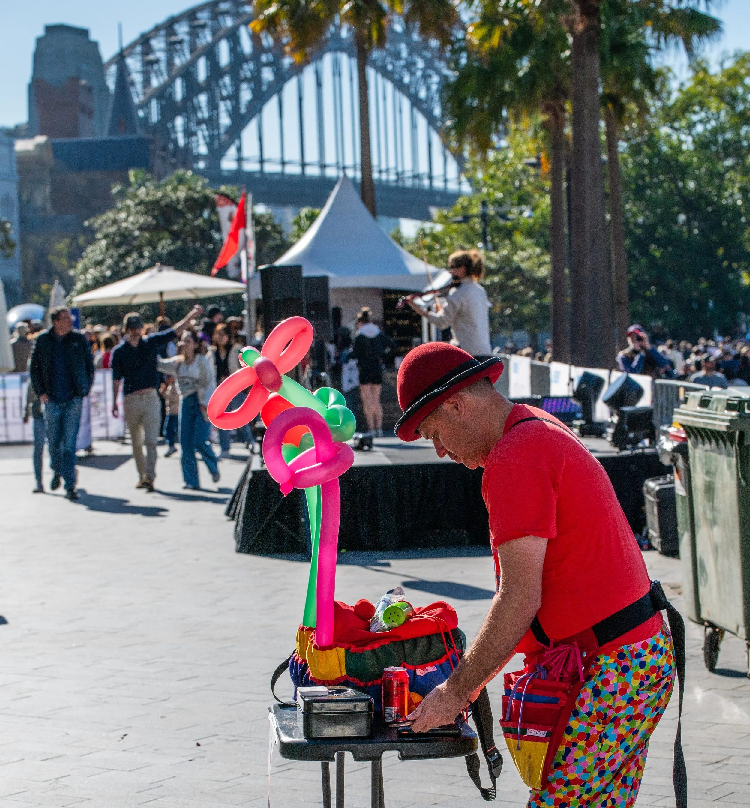 A street performer dressed in colorful, clown-like pants and a red shirt, wearing a red bowler hat, prepares balloon animals on a small table during a busy outdoor event. There are crowds of people in the background, with a stage and a large bridge visible under a clear blue sky.