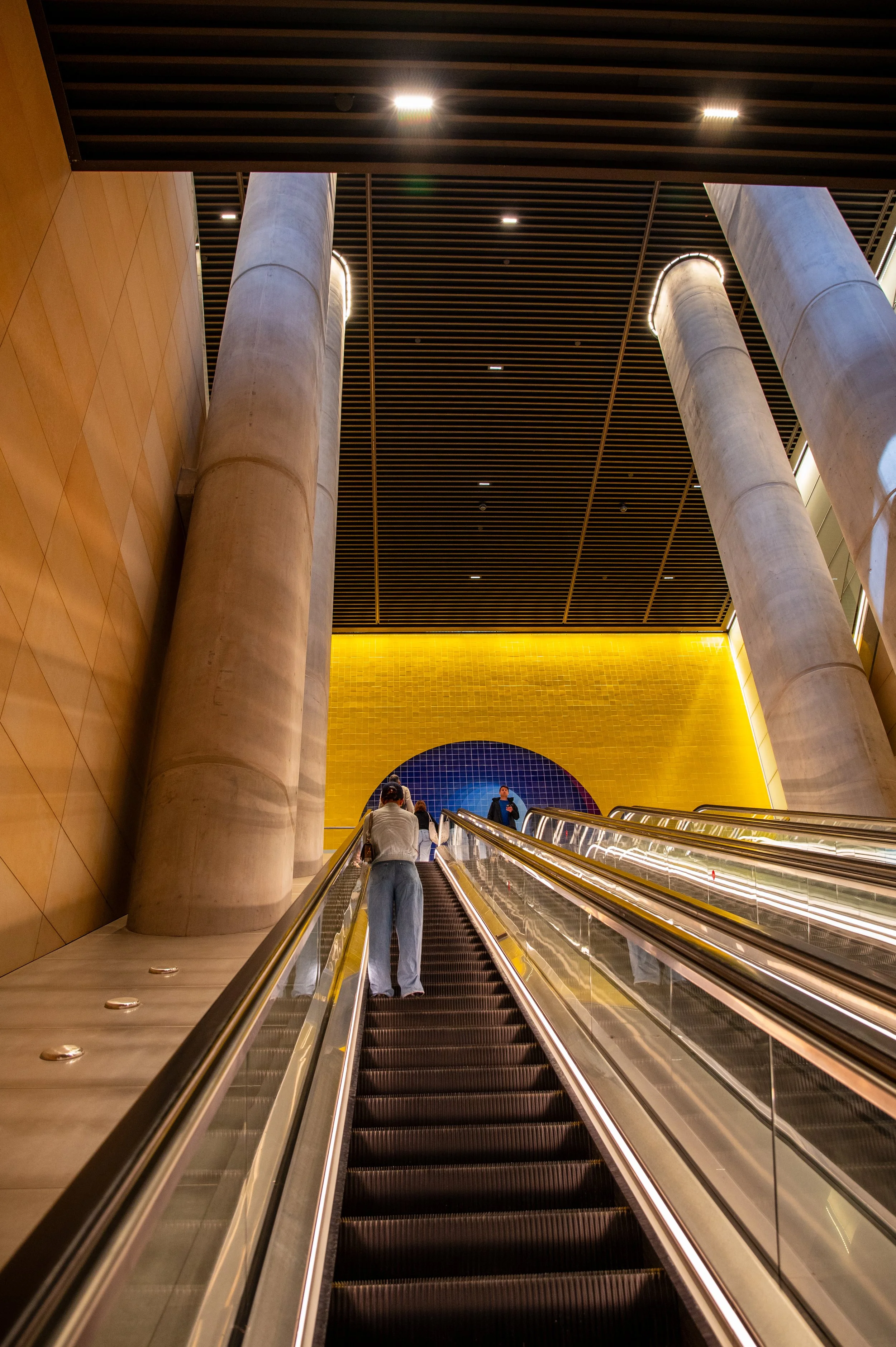 Interior of a building with a tall escalator, large concrete columns, and a colorful wall art featuring a blue semi-circle on a yellow background.