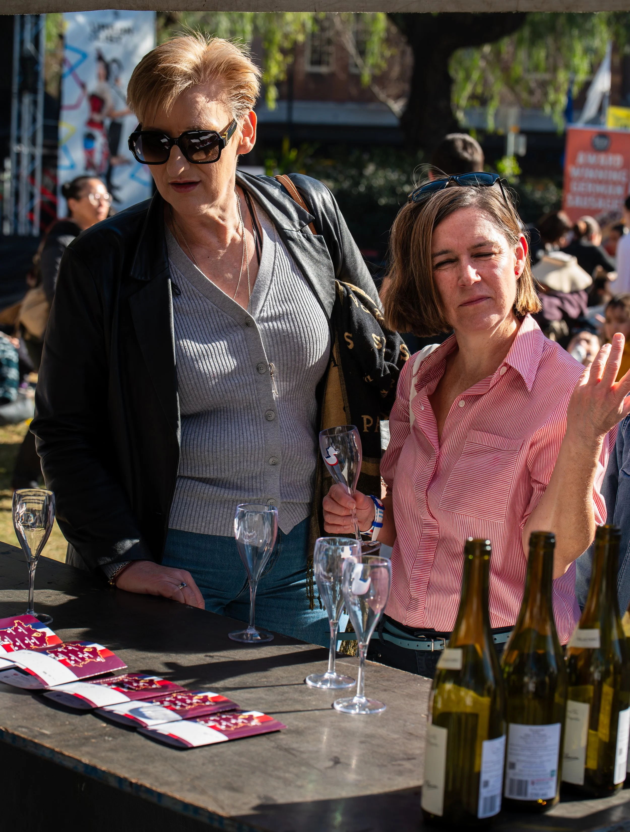 Two women at an outdoor event are standing behind a table with wine bottles, empty wine glasses, and event brochures. One woman with short blonde hair and dark sunglasses is wearing a leather jacket and holding a wine glass; the other with shoulder-length brown hair, striped red and white shirt, is gesturing with her hand.