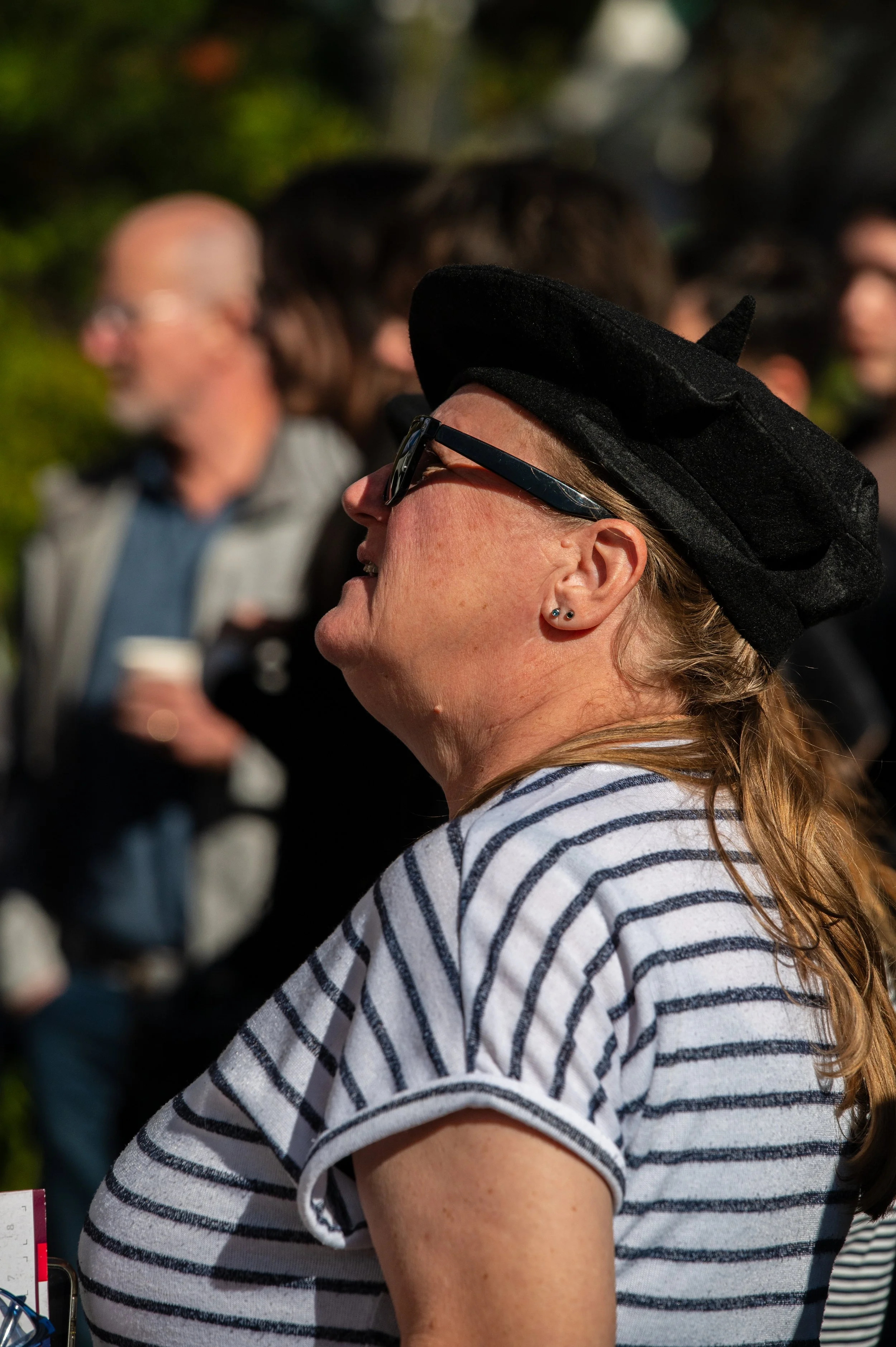 A woman wearing a black hat, sunglasses, and a striped shirt, standing in profile among a crowd of people outdoors.