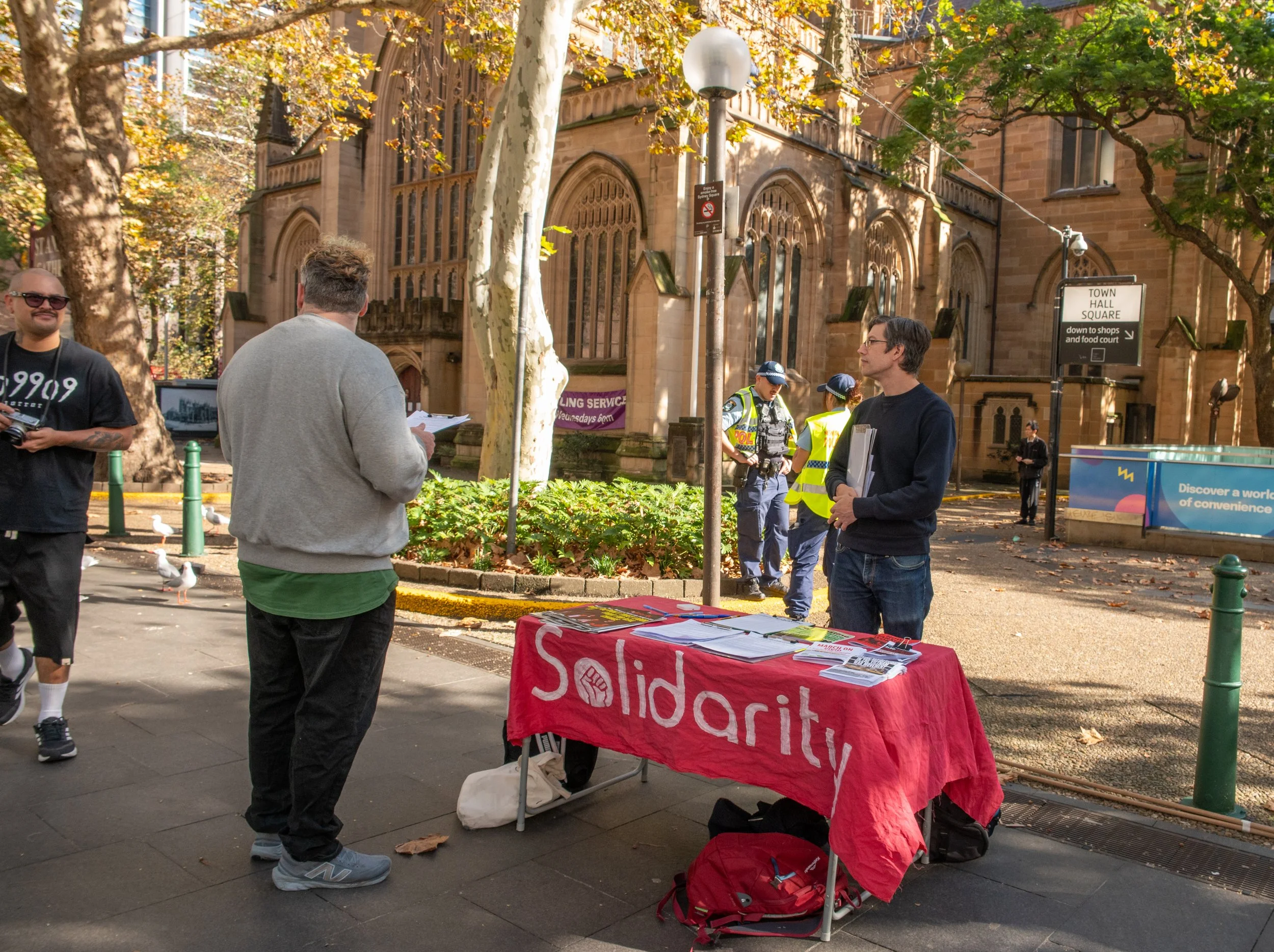 A street scene with a table covered in a red cloth displaying the word 'Solidarity' and people engaged in conversation, with police officers and a church in the background.