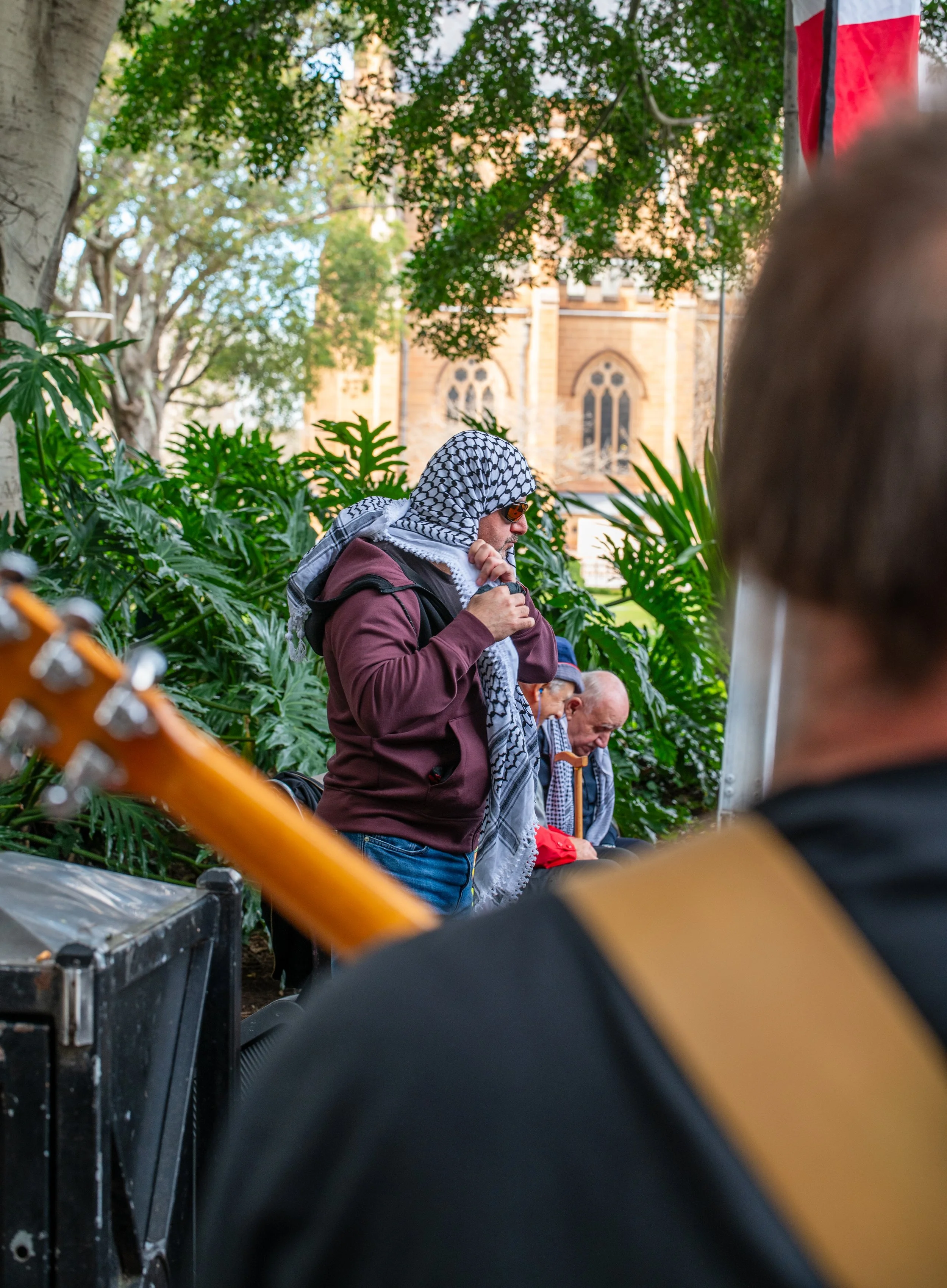 A person wearing a keffiyeh-style scarf, sunglasses, and a burgundy hoodie standing among lush green plants, with others sitting on benches behind. A guitar is partially visible in the foreground.
