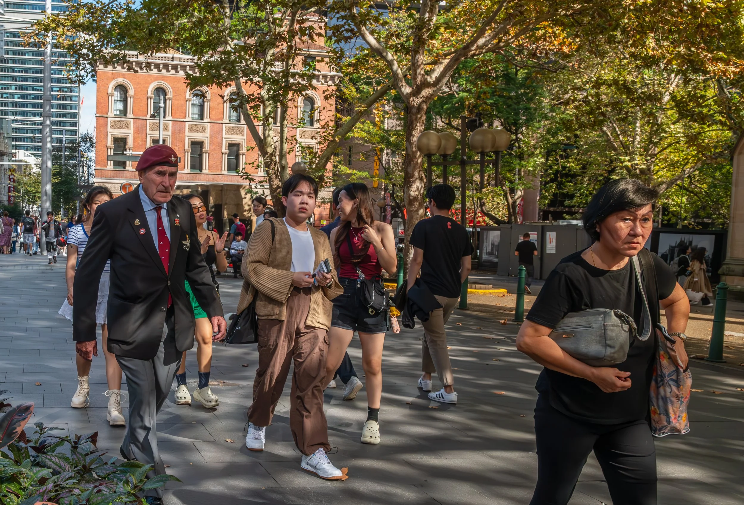 People walking outdoors on a city sidewalk with trees and buildings in the background.