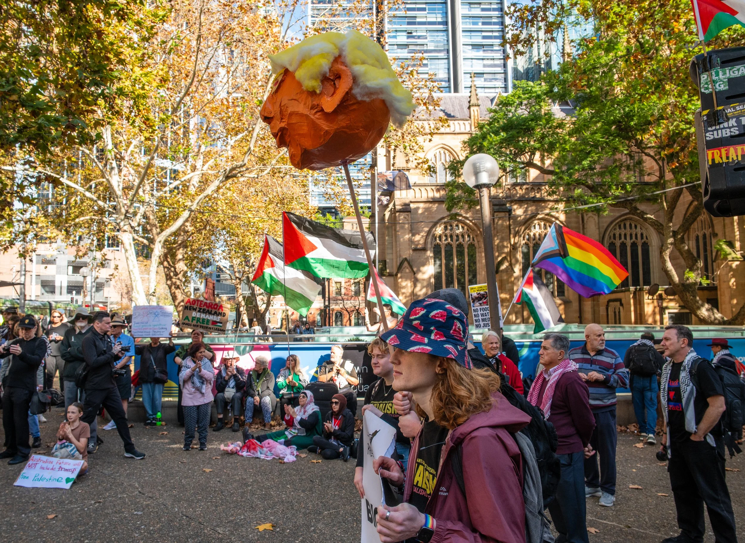 A group of people participating in a protest or rally, holding Palestinian flags, rainbow flags, and signs, with colorful papier-mâché balloon resembling Donald Trump with yellow hair, against a cityscape backdrop with trees and a historic building.
