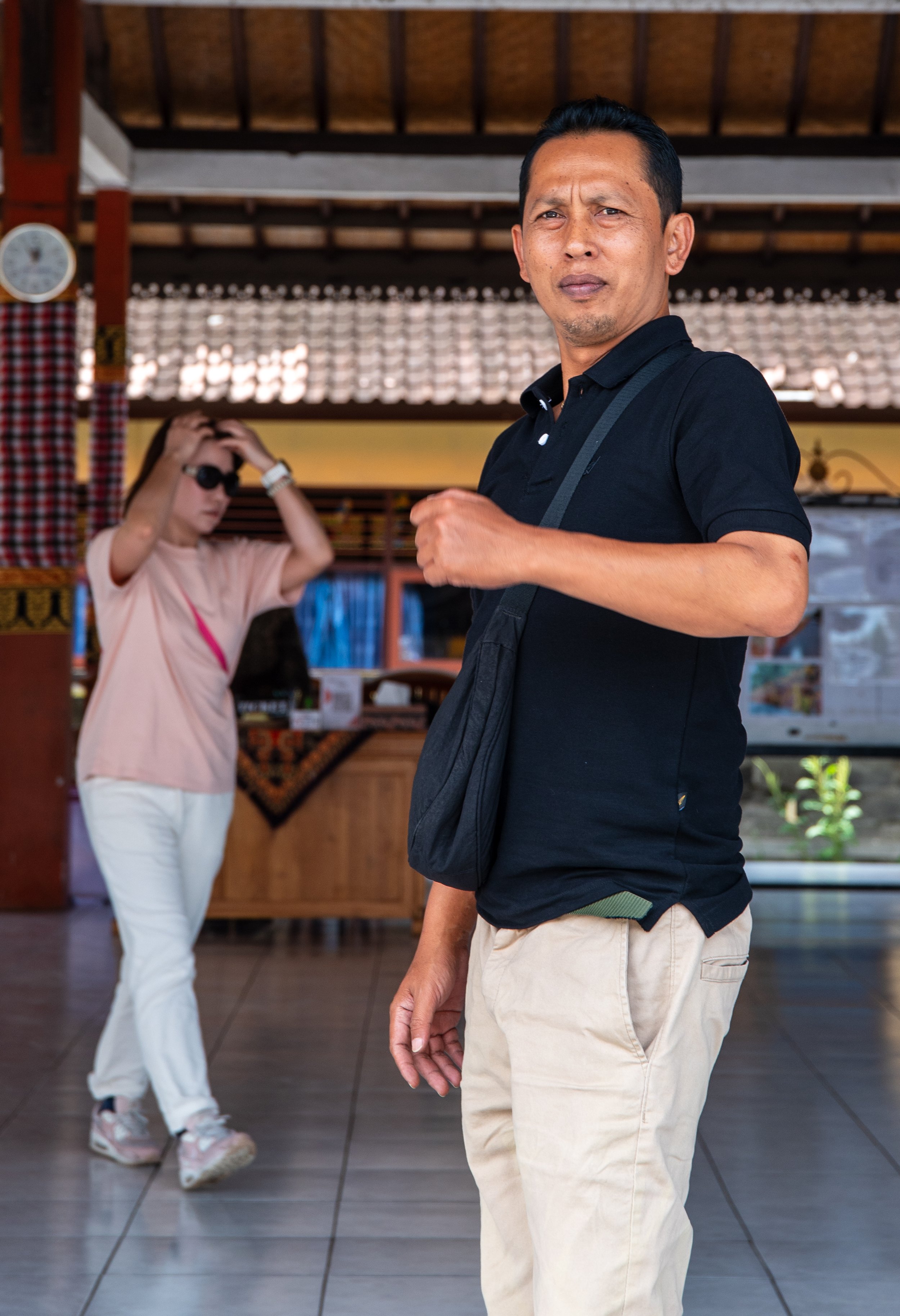 A man wearing a black polo shirt and beige pants stands inside a building, with a woman in the background adjusting her sunglasses and wearing a pink shirt and white pants.