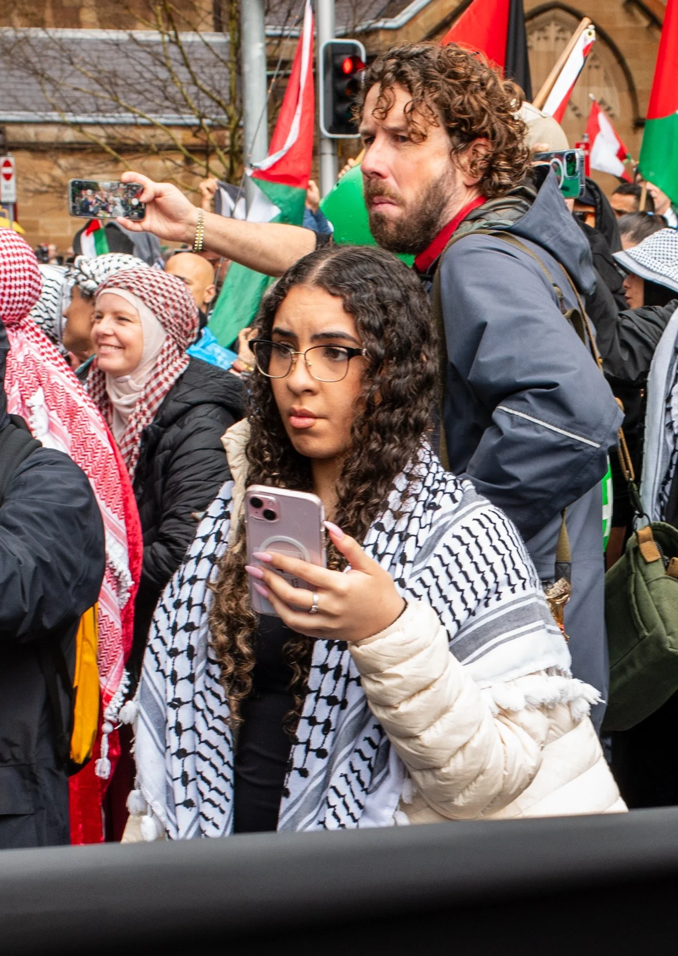 A diverse group of people participating in a protest or demonstration, with some waving flags and taking photos with their phones.