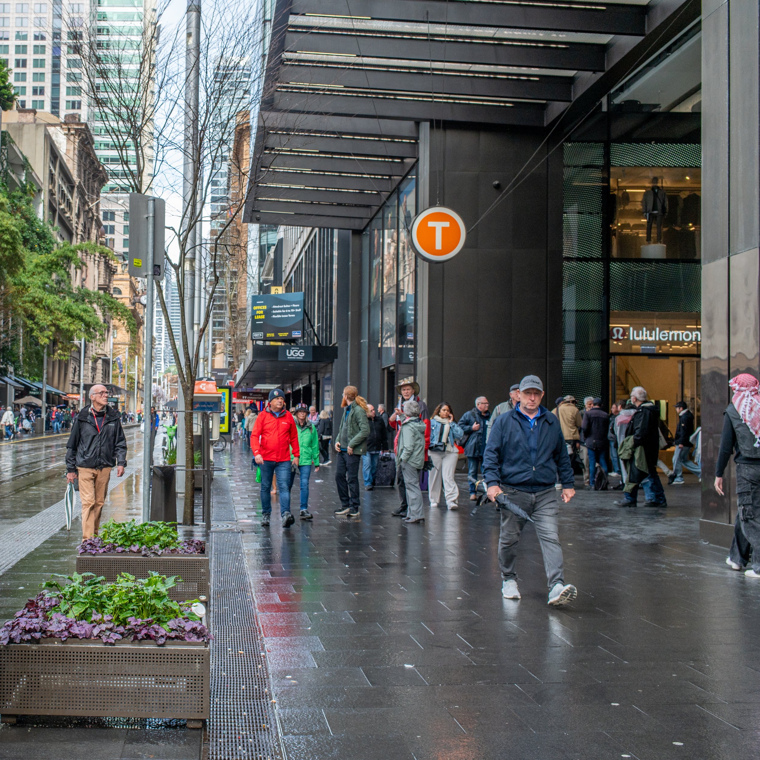 Pedestrians walking past the entrance to a subway station marked with a bright orange 'T' sign on a rainy city street.