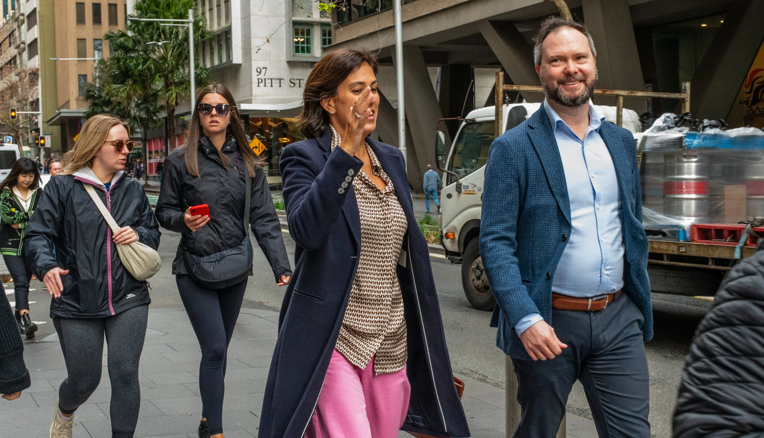 A group of people walking on a city street, including women in athletic wear, and two well-dressed individuals, one man smiling and a woman gesturing.