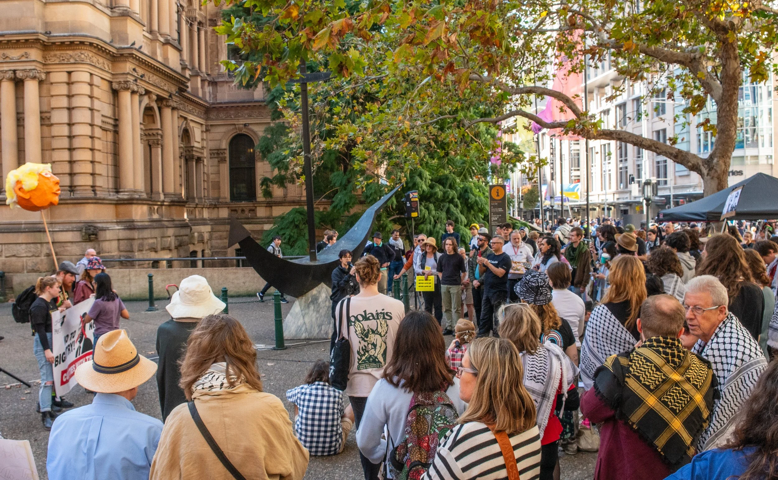 Crowd gathered in an urban park with a modern sculpture and a historic building in the background, some protesters holding signs and a large carved face balloon float.