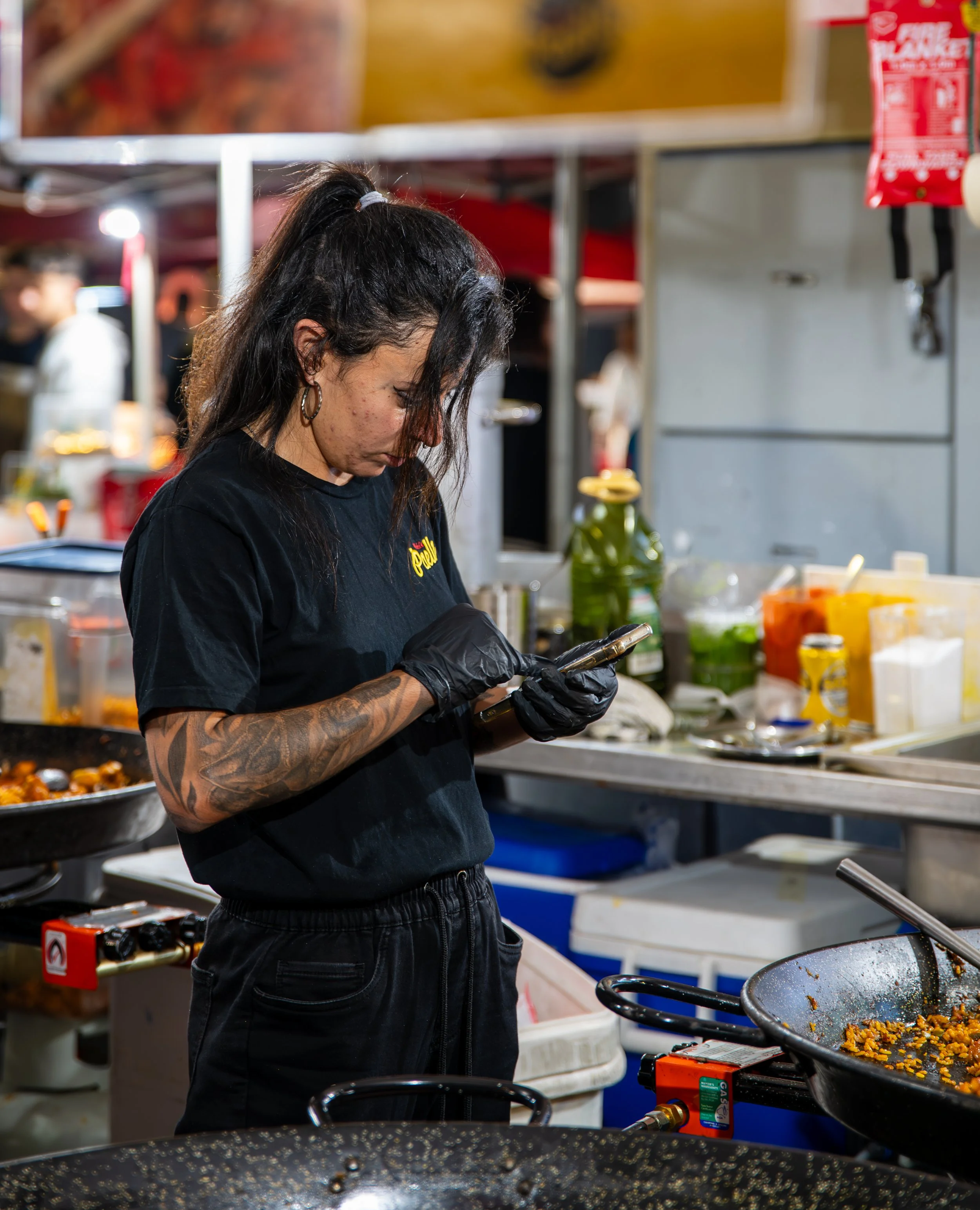 A woman with long dark hair, tattoos on her arm, wearing a black T-shirt and gloves, looking at her phone in a busy outdoor food stall with cooking utensils, ingredients, and condiments around her.
