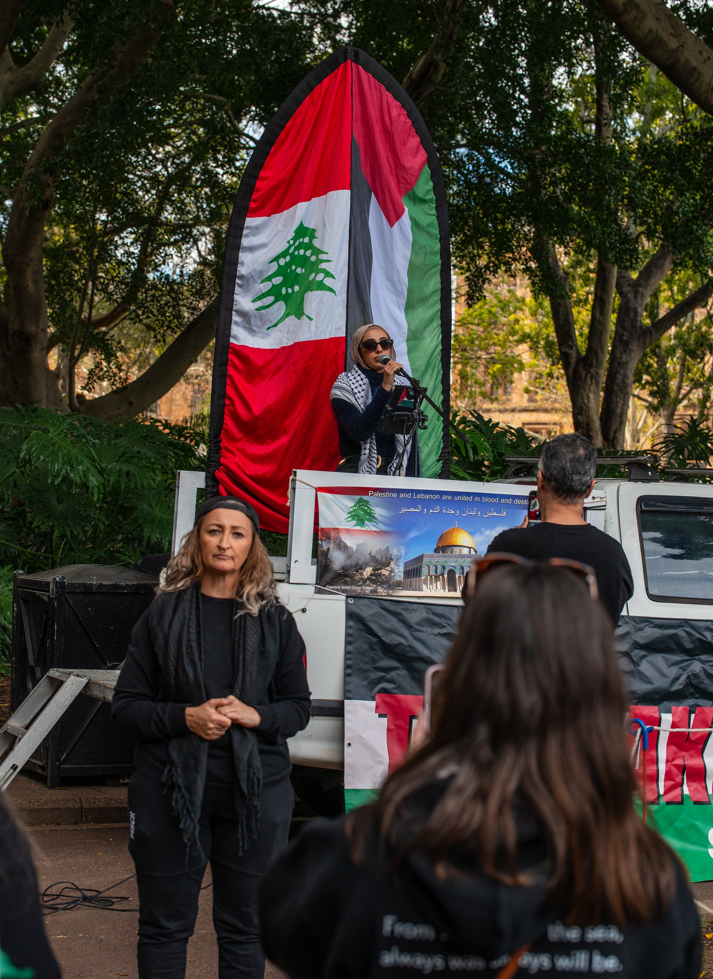 A woman wearing sunglasses and a headscarf speaks into a microphone on a stage with a large Palestinian flag in the background. Several people are listening, and there is a sign with images of Palestinian landmarks and a message about Palestine and Lebanon in the foreground.