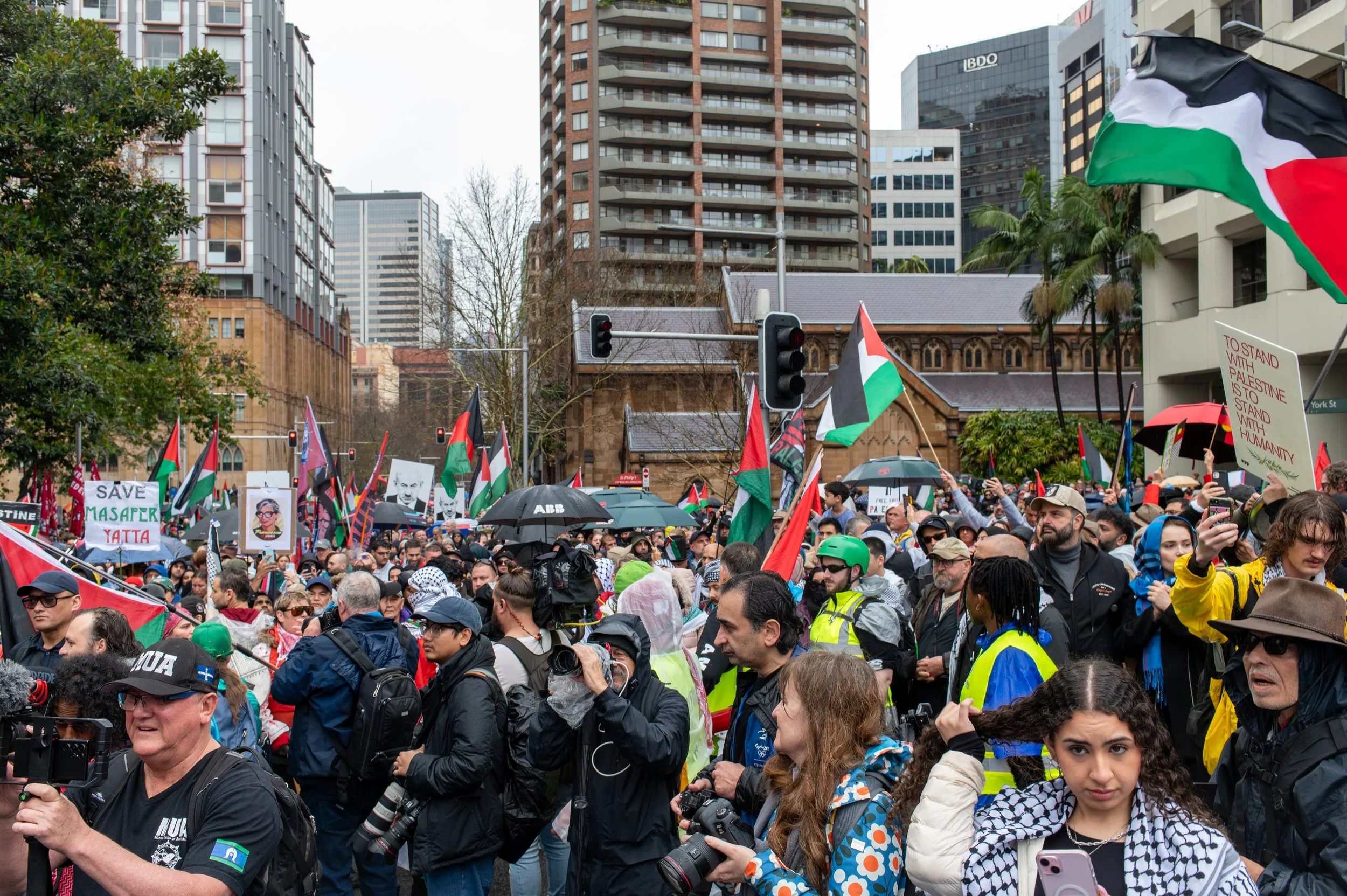Crowd of protesters holding Palestinian flags, signs, and banners at a demonstration in an urban city setting with high-rise buildings in the background.