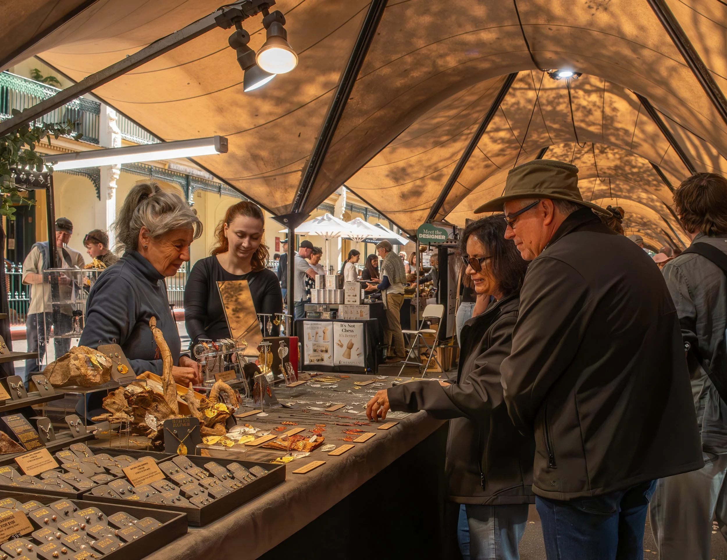 People shopping at an outdoor jewelry market stall, browsing gold and gemstone jewelry items under a canopy, with a few vendors and other shoppers visible in the background.