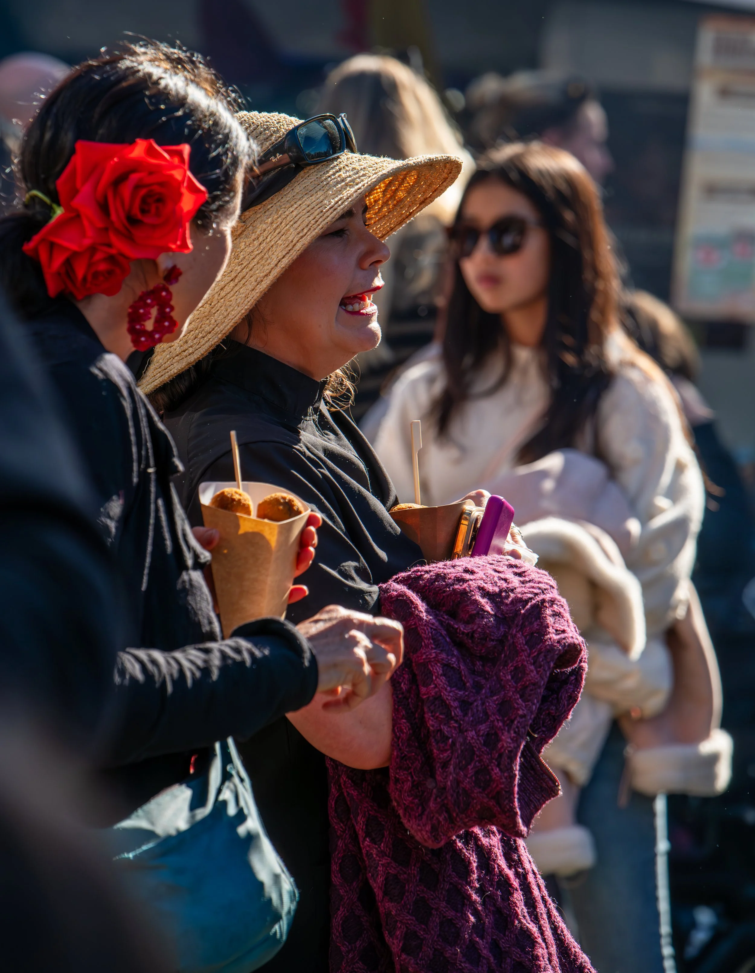 Three women talking outdoors, one wearing a large straw hat and sunglasses, holding food, and another with a red flower in her hair, holding a brown paper container. The background has other people and blurry signs.