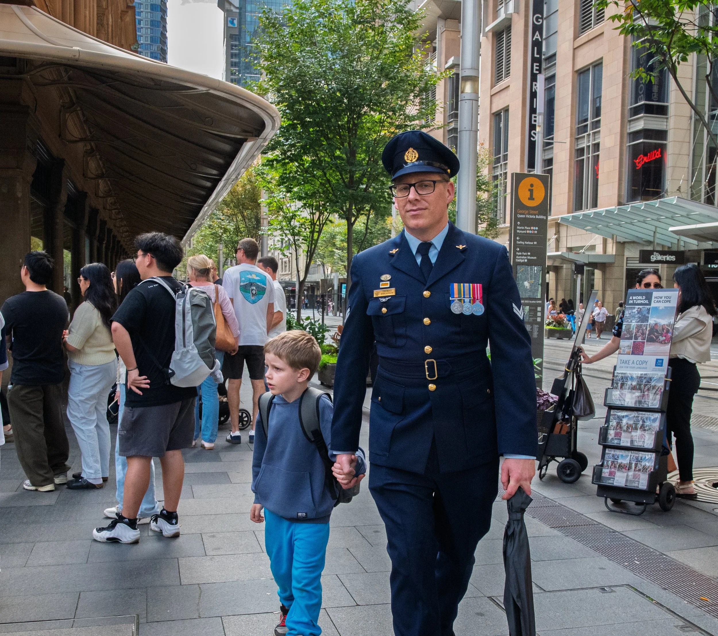 A police officer in a formal uniform walking hand in hand with a young boy on a busy city sidewalk, with people waiting in line and tall buildings around.