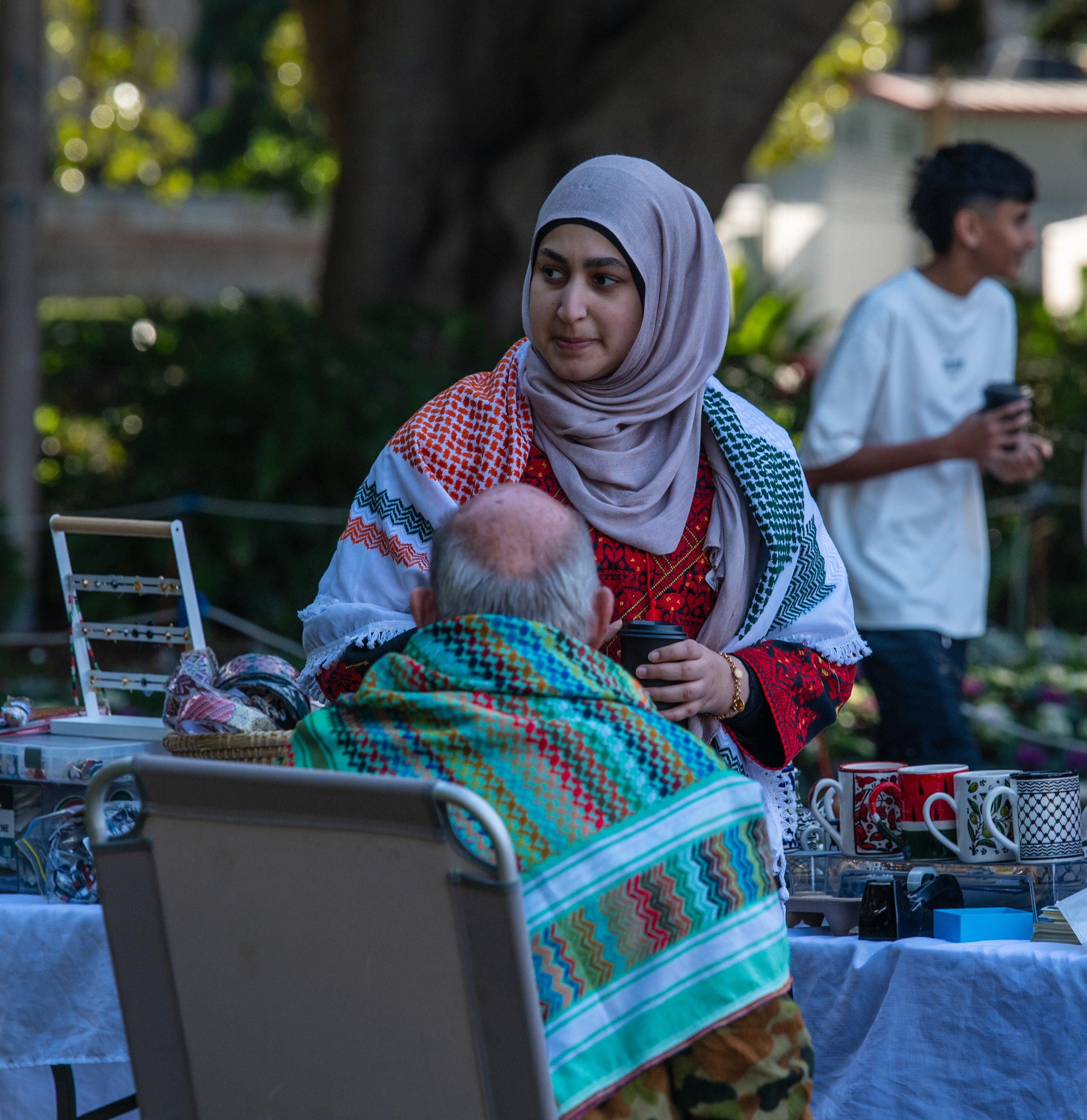 A young woman wearing a hijab and colorful traditional clothing is talking to an elderly man wrapped in a vibrant scarf at an outdoor table with mugs and other items. A young man is in the background using his phone in a park setting with trees.