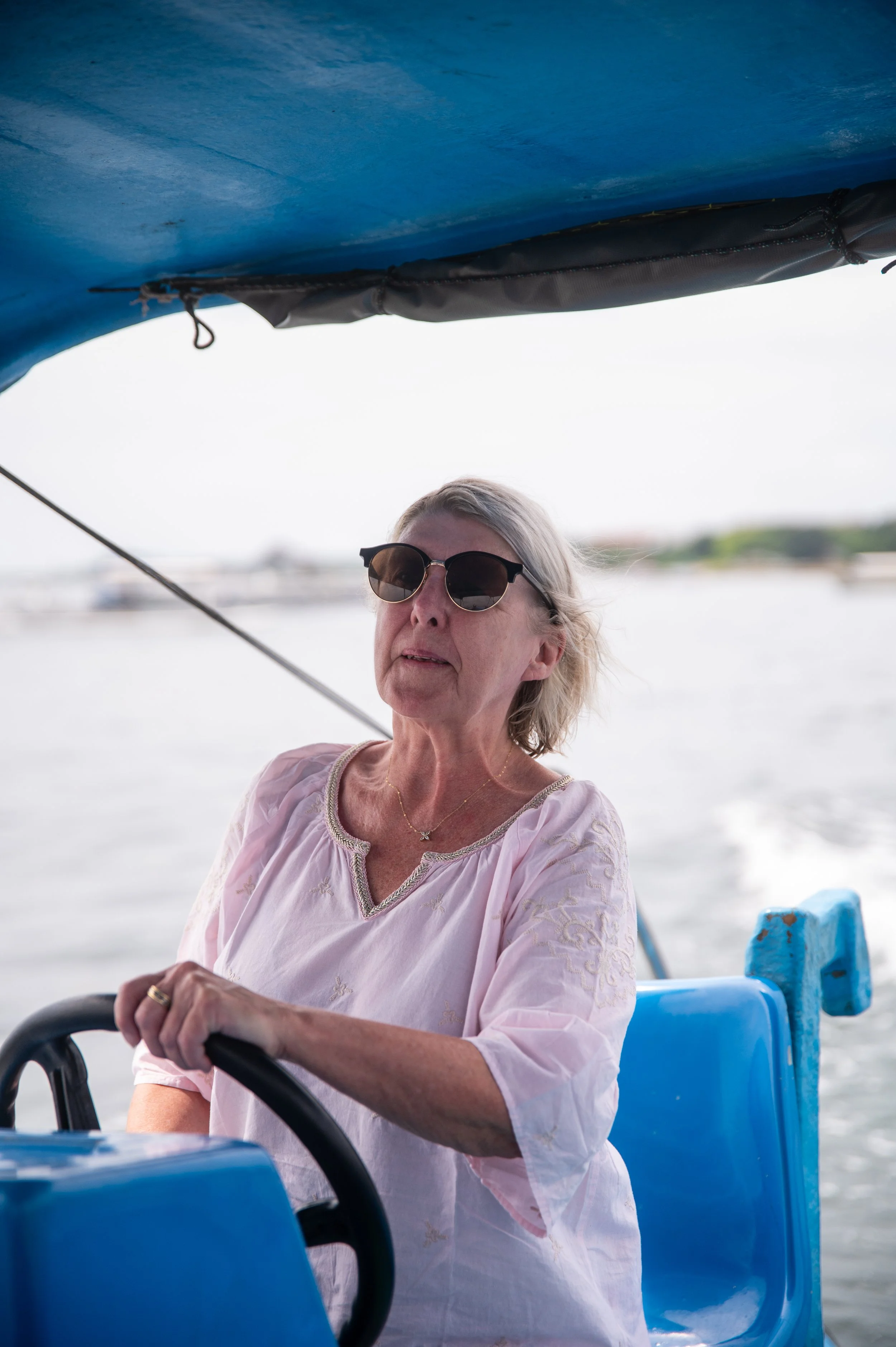 A woman with gray hair, wearing sunglasses and a pink embroidered blouse, is sitting on a boat and holding a steering wheel, with water in the background.