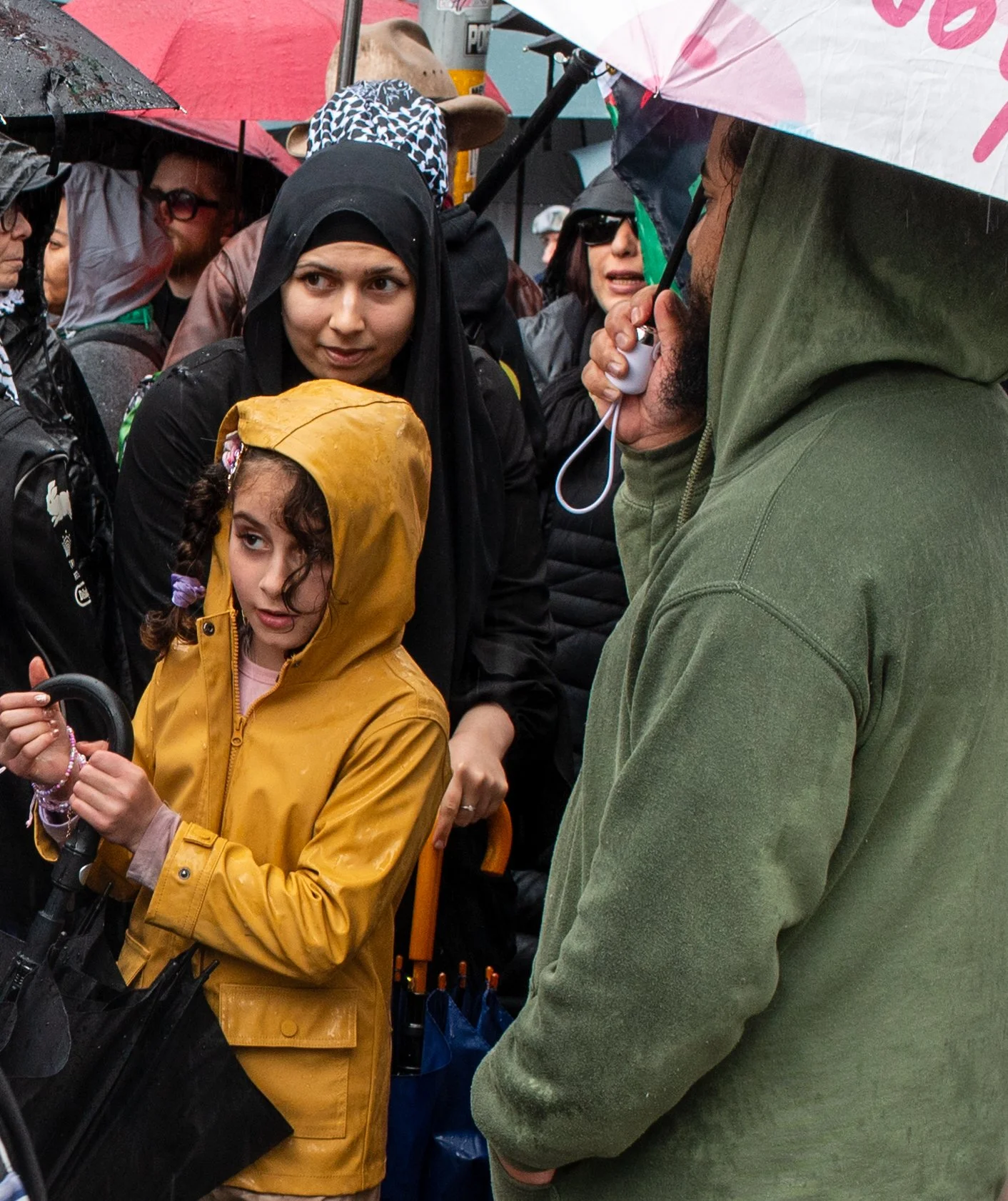 A group of people gathered outdoors in rainy weather, some holding umbrellas. A young girl in a yellow raincoat stands in front, holding an umbrella handle. A woman next to her watches attentively. A man in a green hoodie is speaking into a microphone or megaphone, partially visible on the right. The scene appears to be at a protest or rally.