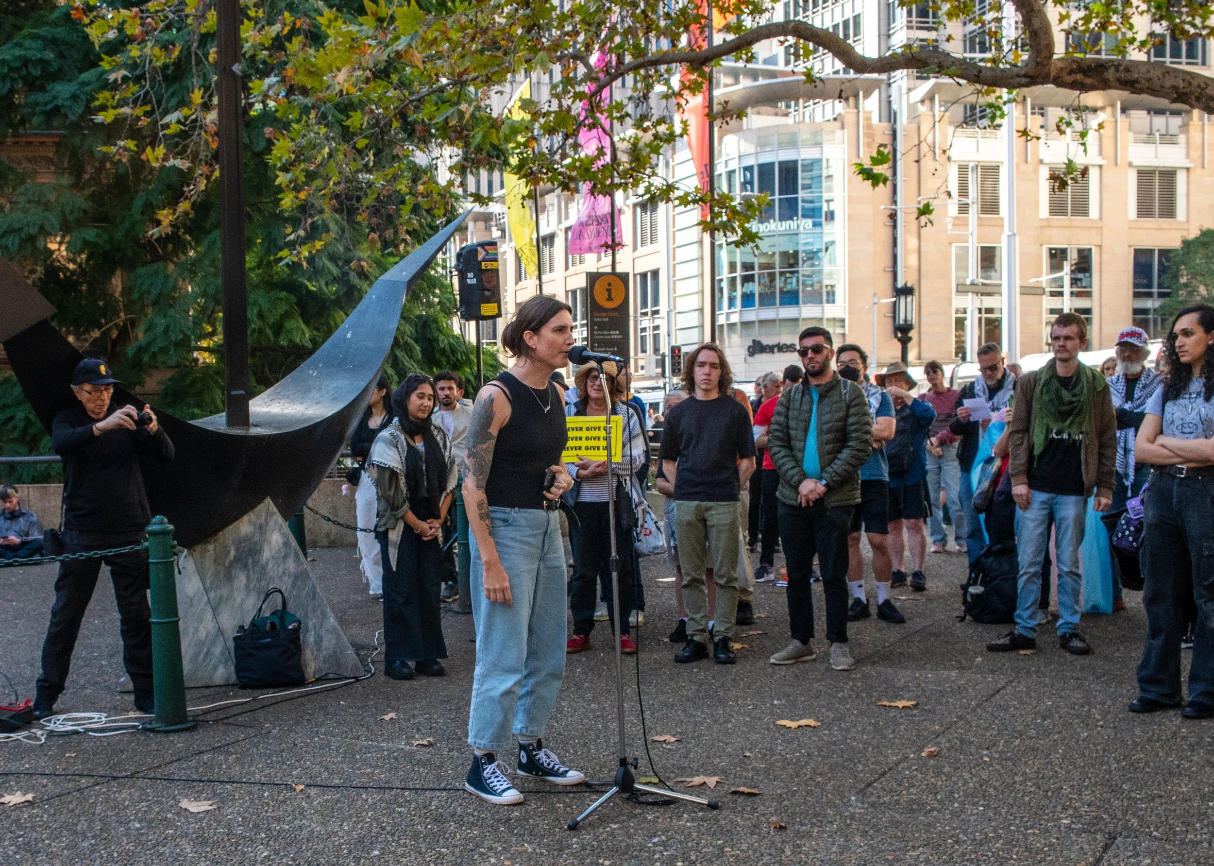 A woman with tattoos and black tank top speaking at a microphone during a public gathering in an urban park, surrounded by a diverse group of people, trees, and city buildings.