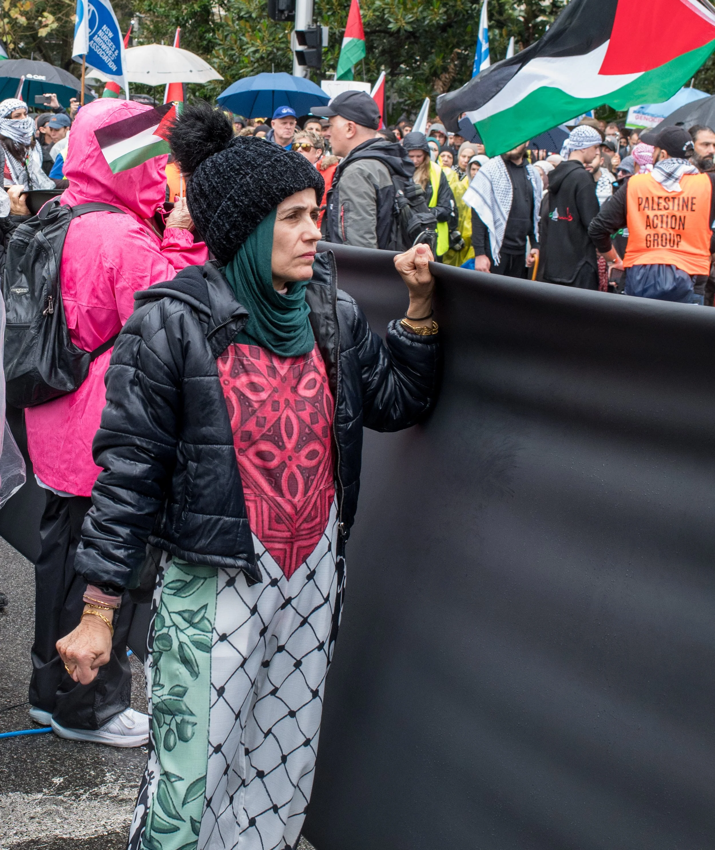 A woman holding a black banner at a protest or rally, with a crowd of people, many waving Palestinian flags, in the background.