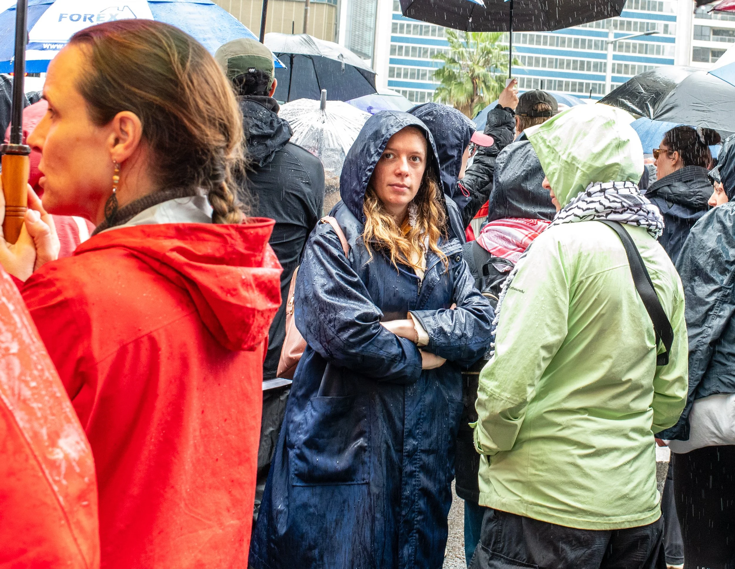 A group of people gathered outdoors in rainy weather, many wearing rain jackets and holding umbrellas. One woman in a dark blue raincoat stands with her arms crossed, looking directly at the camera. Others are visible in the background, some with hoods or umbrellas, against a backdrop of tall buildings.