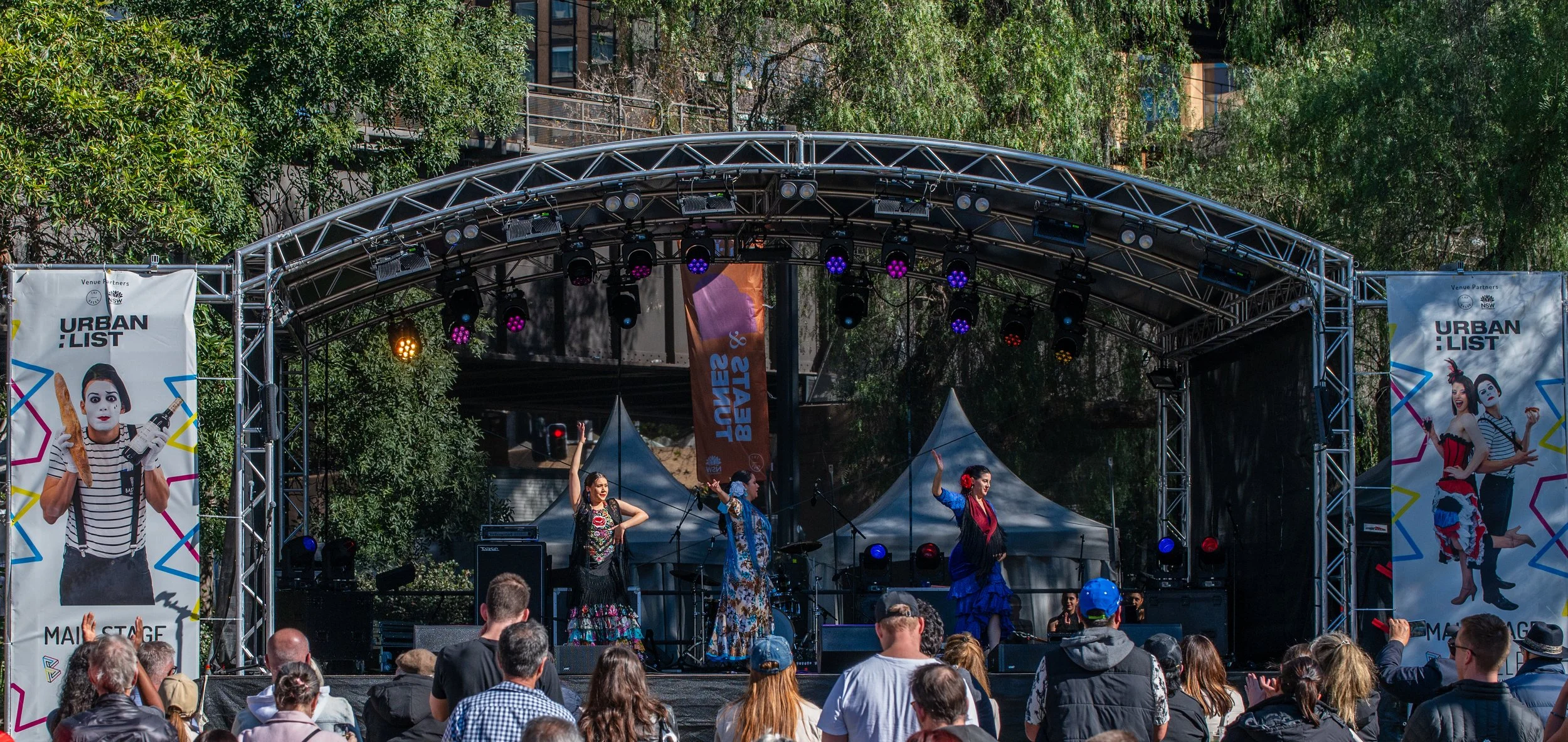 Outdoor stage with performers and audience, event banners, trees, and sky