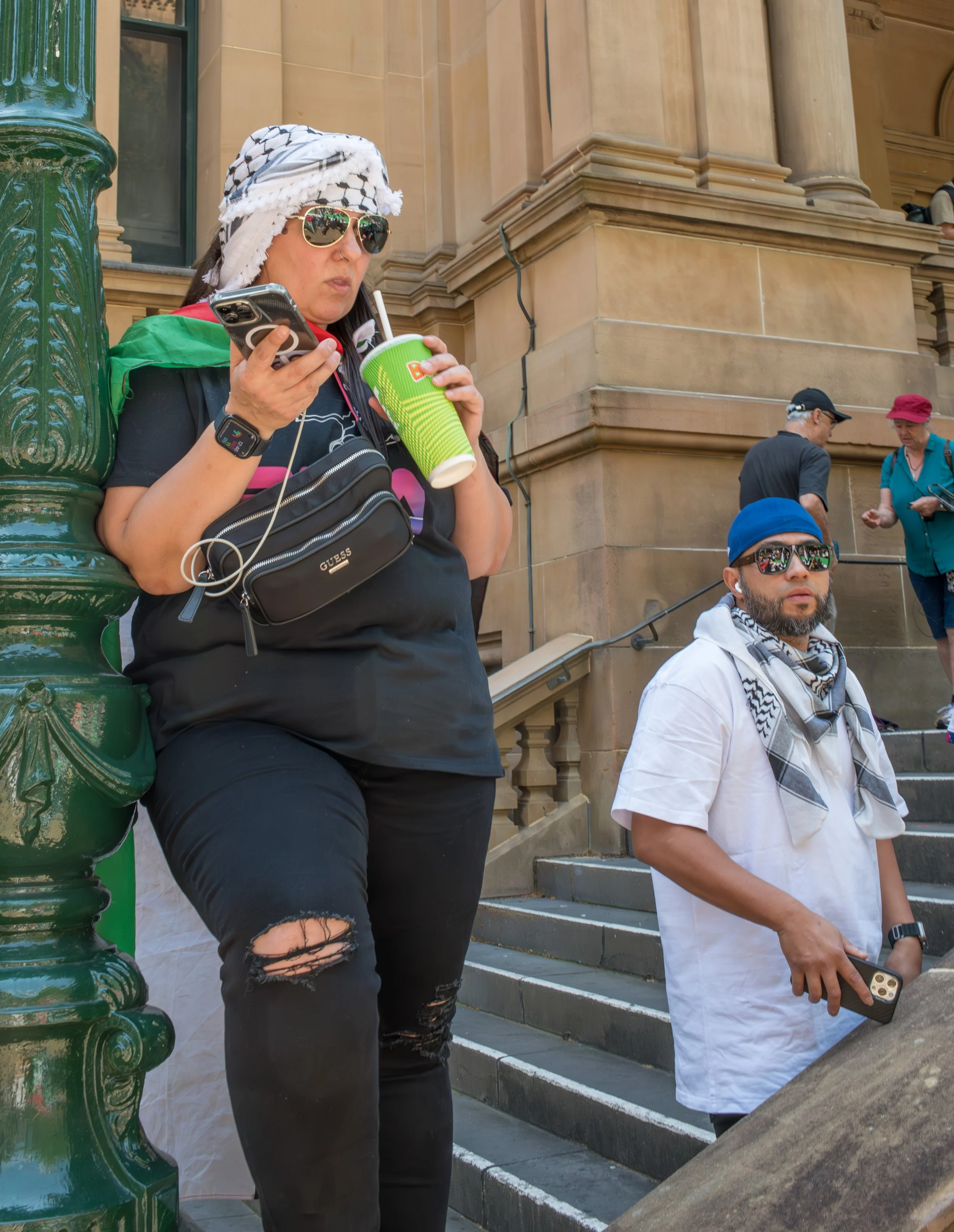A woman wearing sunglasses, a black shirt, and a headscarf drinks from a green cup while looking at her phone, standing near a green lamppost outside a stone building. A man with sunglasses and a blue head covering is nearby, holding his phone, with other people in the background walking on the stairs.