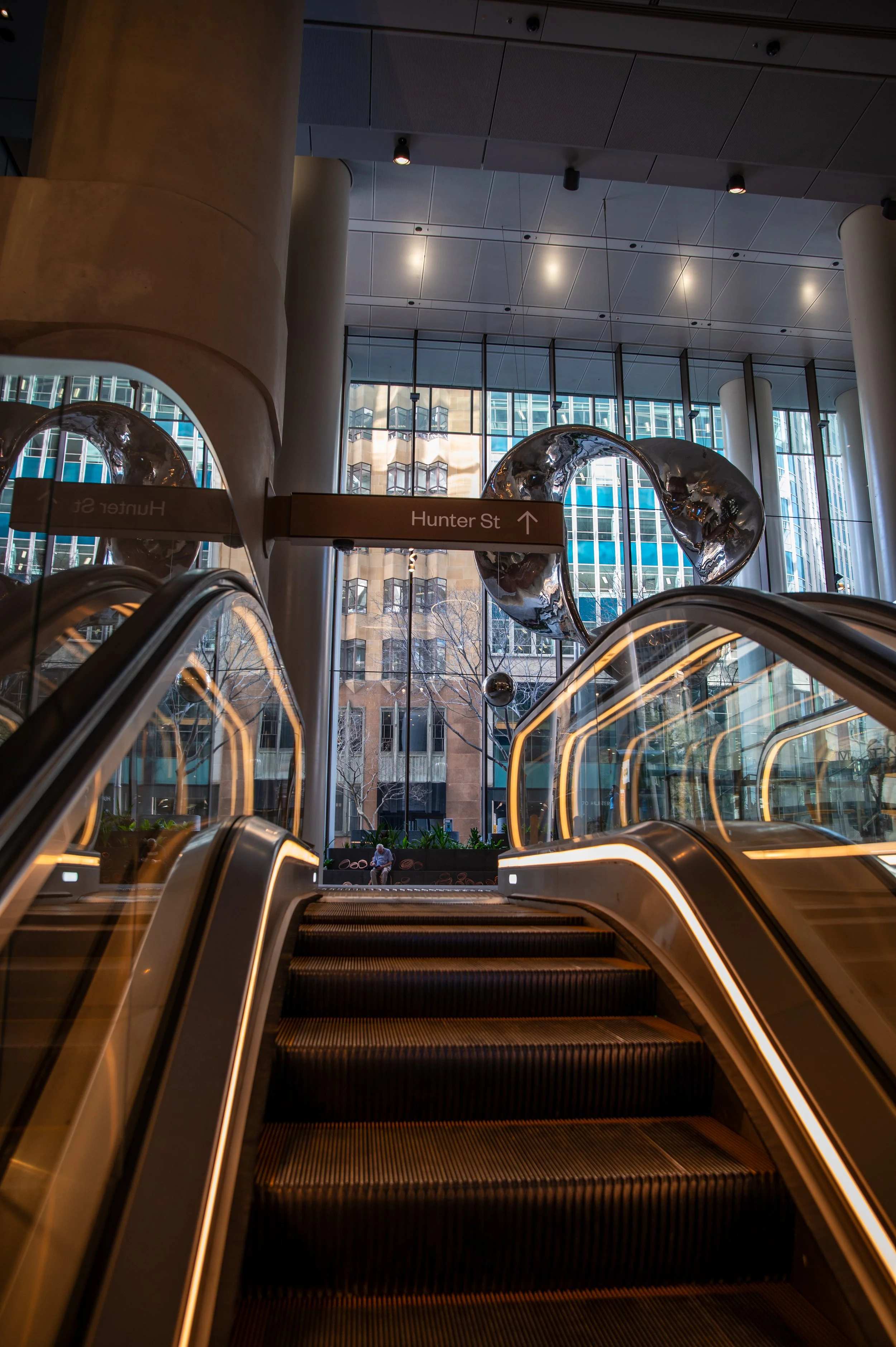 Indoor escalator leading upward in a modern building with large window view of city and reflective art sculptures.
