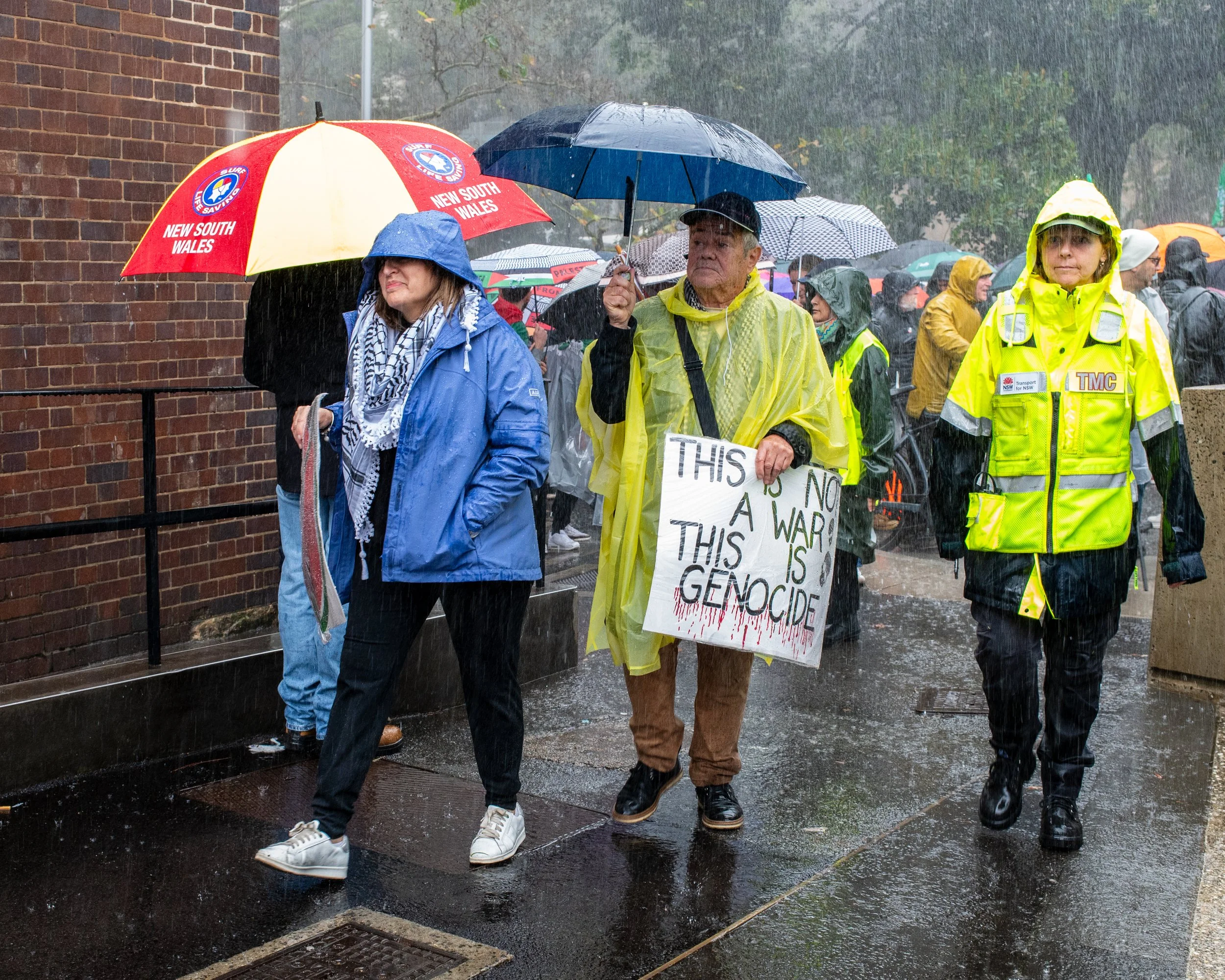 People protesting in the rain, holding umbrellas and wearing rain jackets. One protester holds a sign that says, "This is no war, this is genocide."