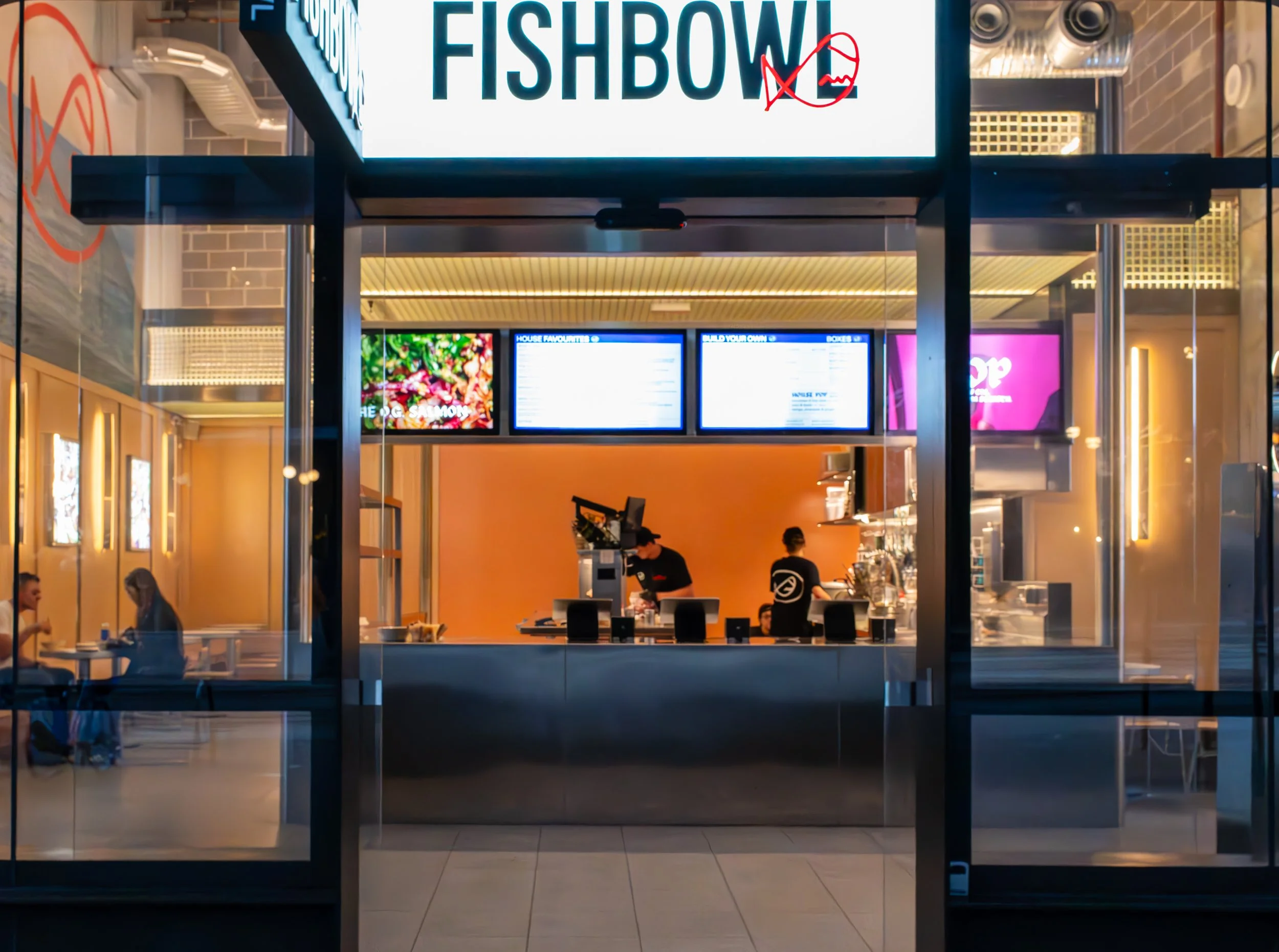 View of a food restaurant named Fishbowl seen from outside, with a sign showing a fish skeleton and a crossed-out fish symbol above the counter. Inside, employees are working behind the counter, and digital menus are displayed above them. There are a few customers seated on the left side.