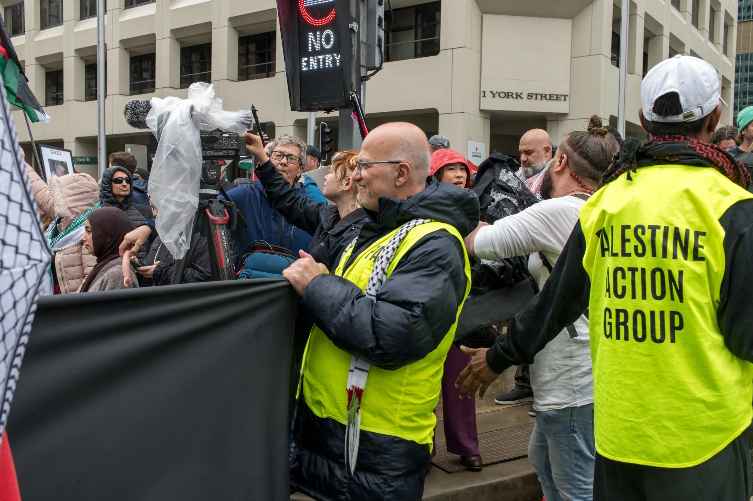 Protesters and media presence at a rally outside a building with a sign reading '1 York Street'. Some protesters are wearing reflective vests, one with 'Palestine Action Group' printed on the back. A camera operator records the scene.