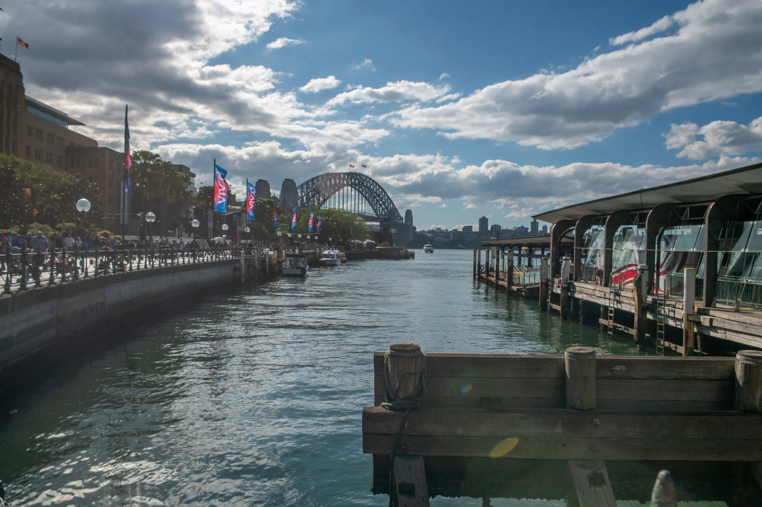 View of Sydney Harbour with the Sydney Harbour Bridge in the background, boats on the water, and a waterfront walkway with flags and people.