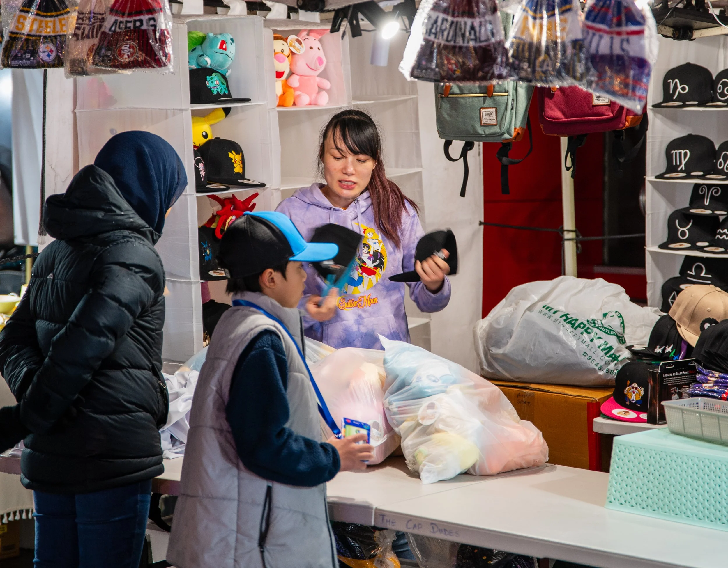 A young woman selling hats and plush toys at a market stall with two children browsing items. The stall has various hats, plush toys, and backpacks on display.