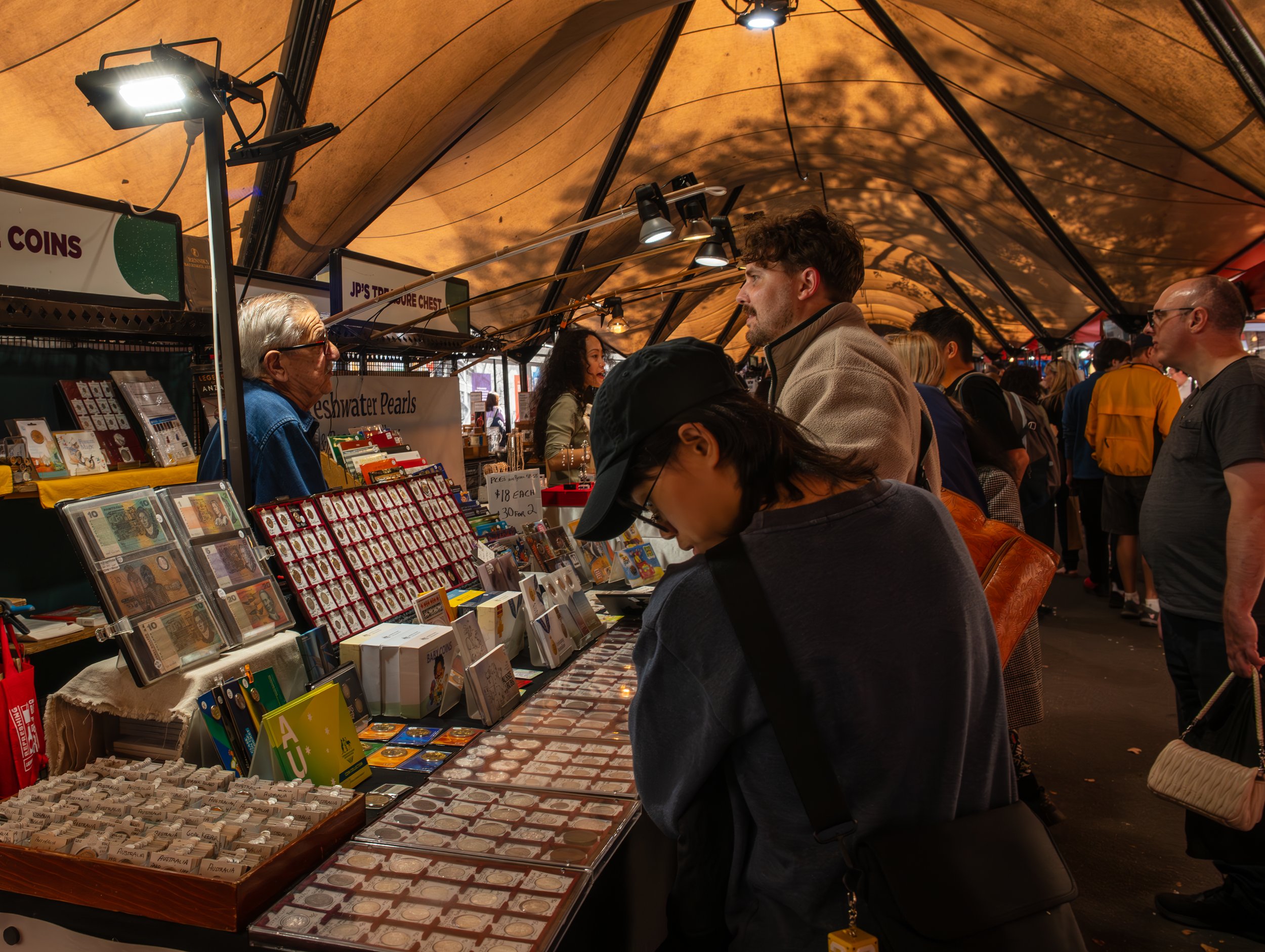 People browsing a marketplace stall with jewelry and collectibles under a canopy, with other shoppers in line and signage for coins and treasure chest jewelry.