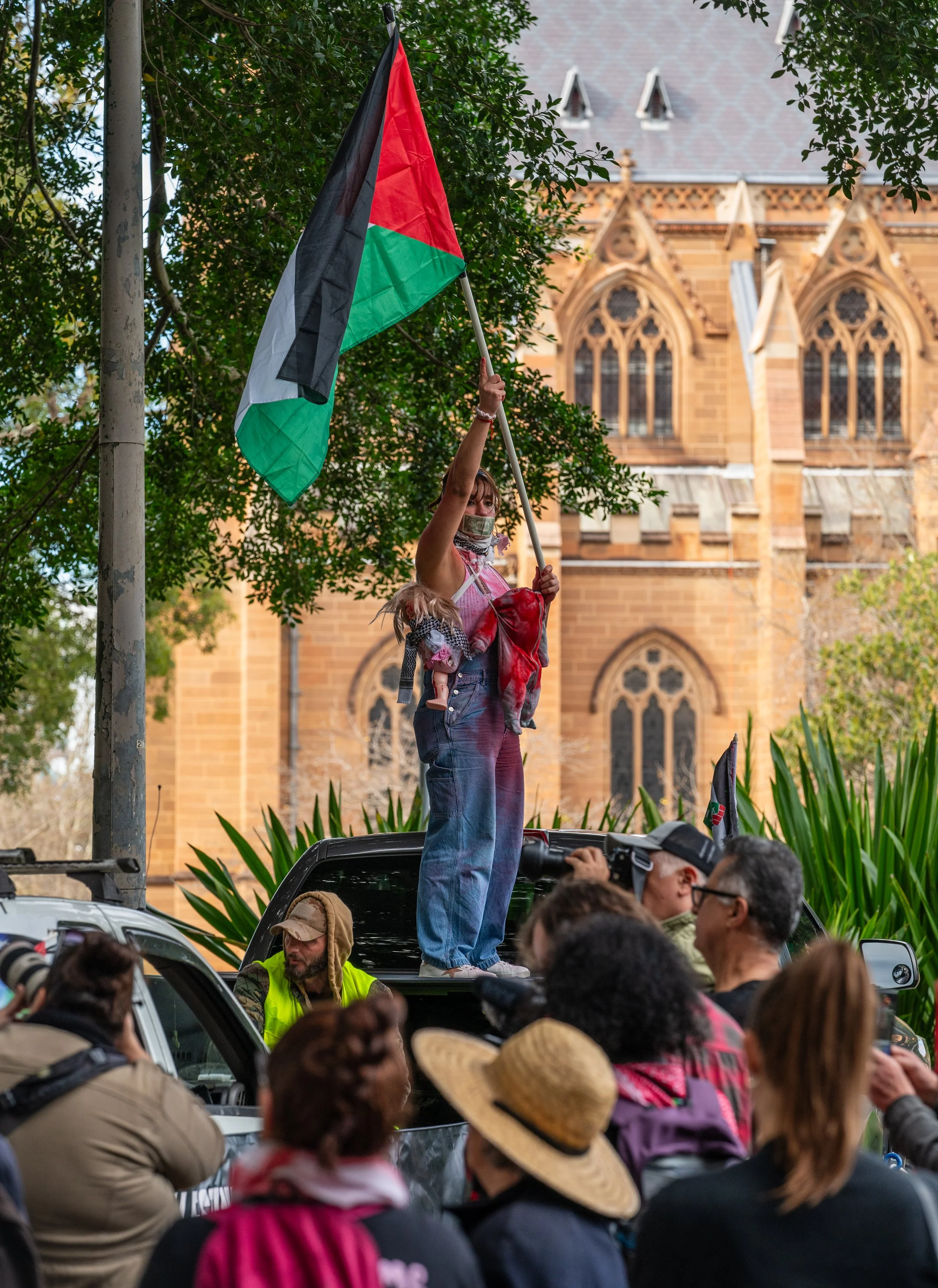 A woman holding a Palestinian flag stands on the back of a vehicle during a protest, surrounded by a crowd. A historic church building is visible in the background.