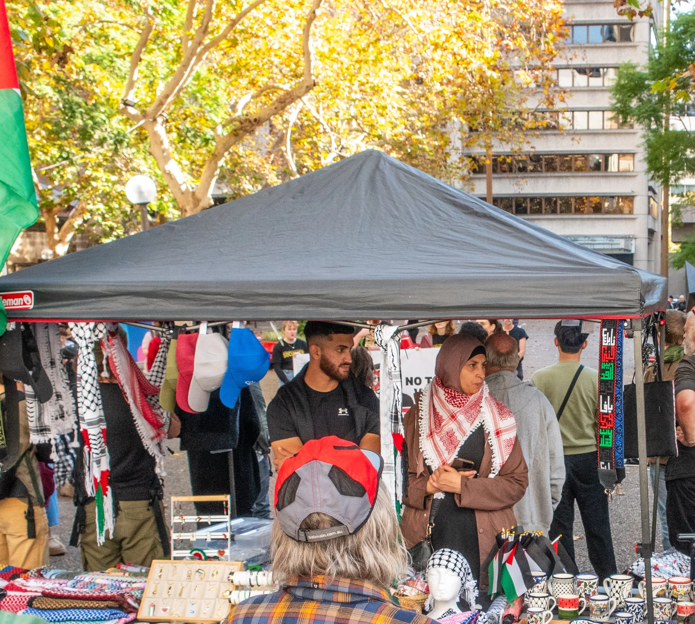 People browsing Palestinian-themed merchandise at an outdoor market stall with a black canopy, surrounded by trees with autumn foliage.