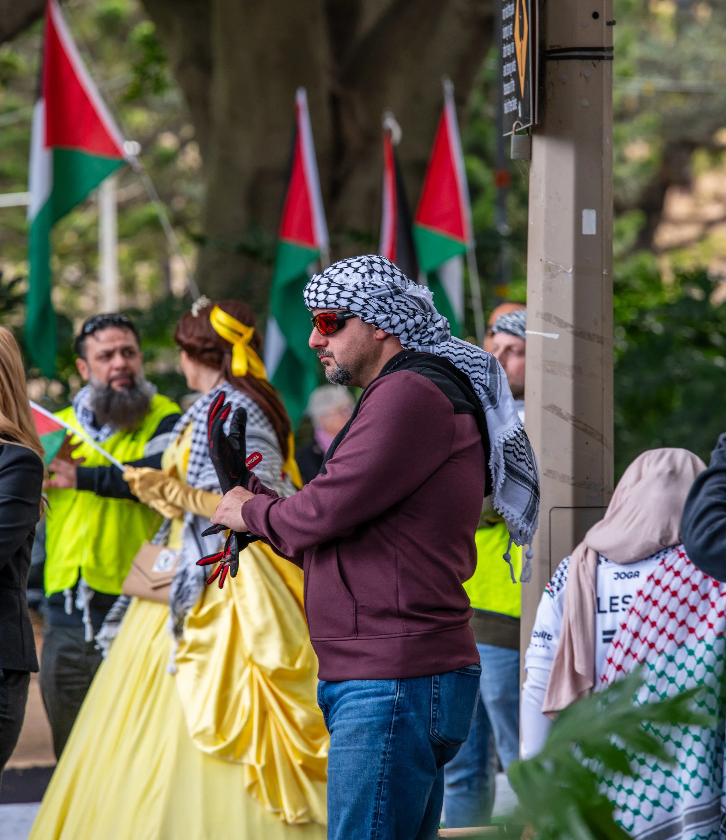 Group of people at a protest or demonstration, some holding Palestinian flags. One man in the foreground wearing a keffiyeh and sunglasses is holding gloves. Other individuals are dressed in yellow and black, with some wearing Palestinian keffiyeh scarves as well.