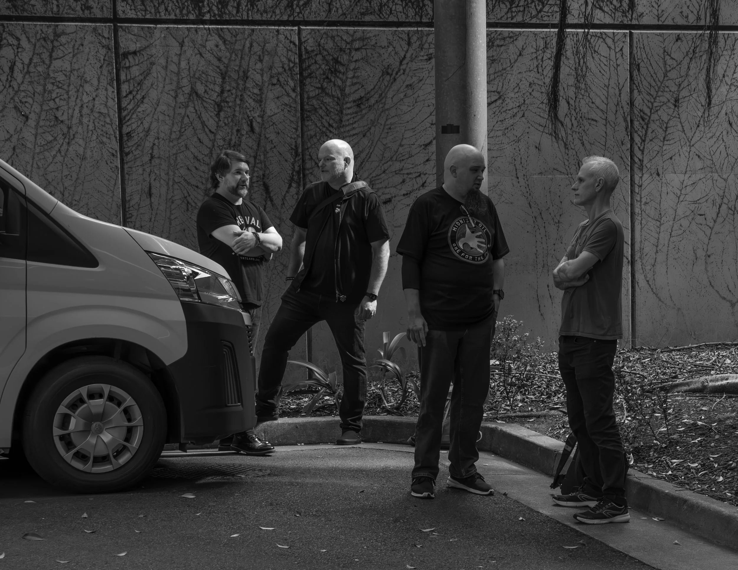Four men standing and talking near a parked vehicle in an outdoor setting with a painted wall background.