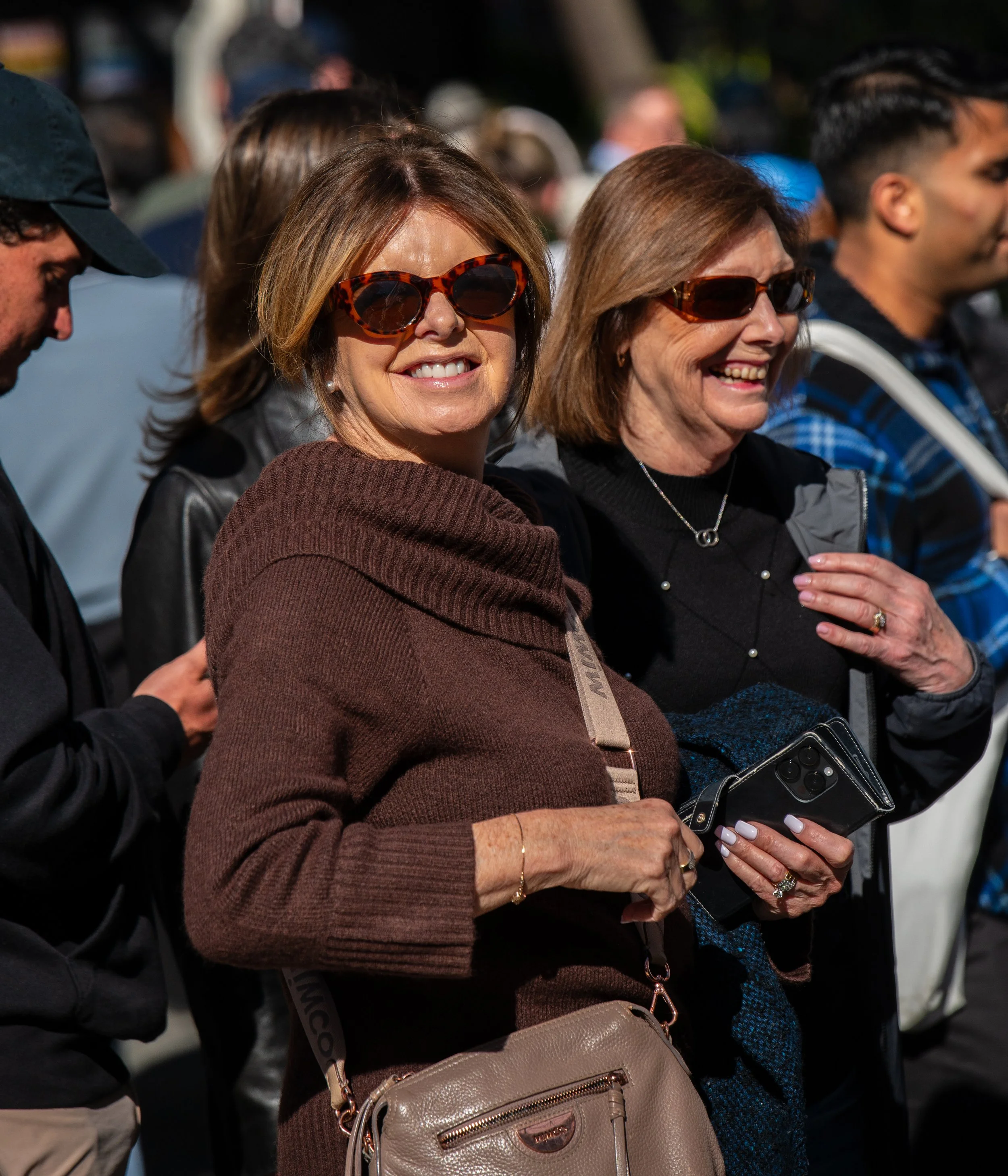 Two women with sunglasses smiling at an outdoor event, one holding a smartphone, surrounded by other people.