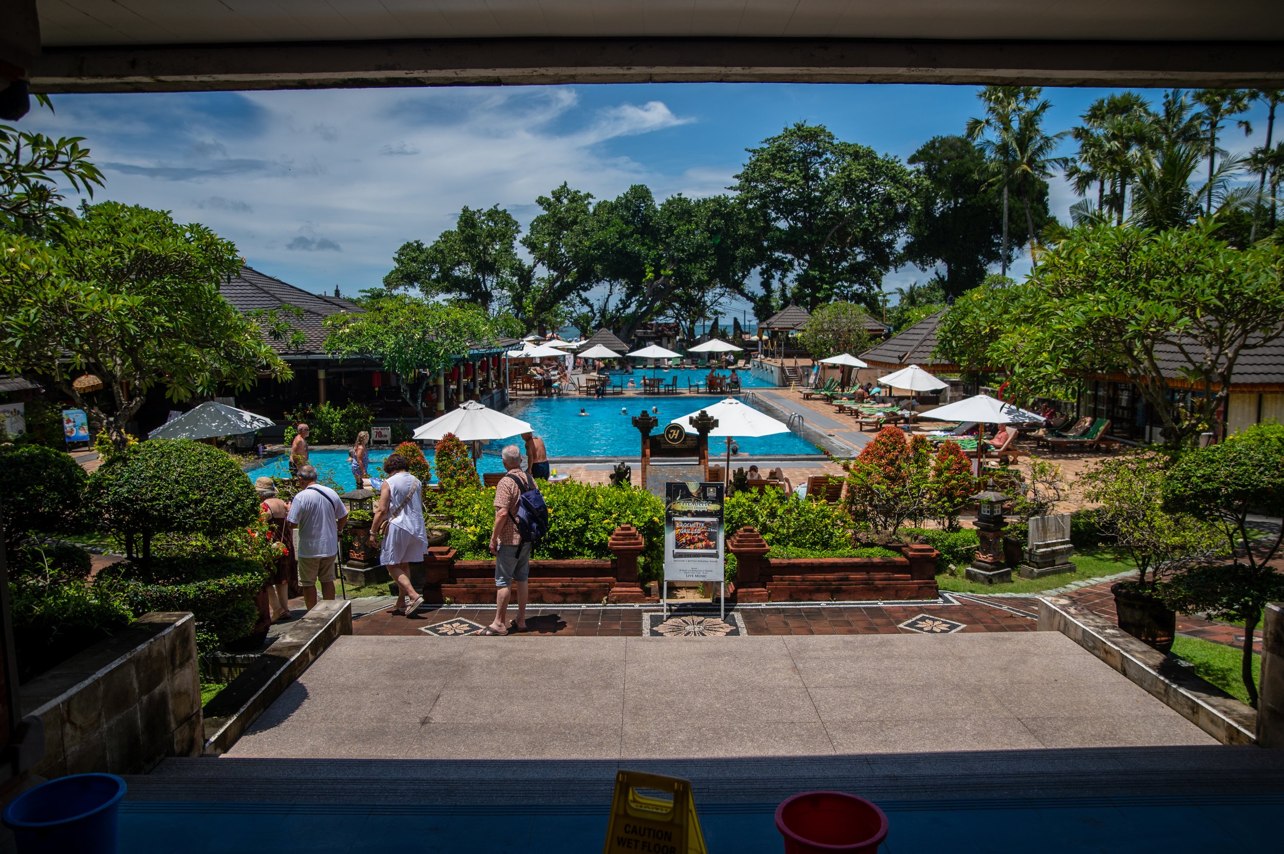 View from inside a building looking out at a busy outdoor swimming pool area with people swimming and relaxing under umbrellas, surrounded by lush green trees and tropical landscaping.