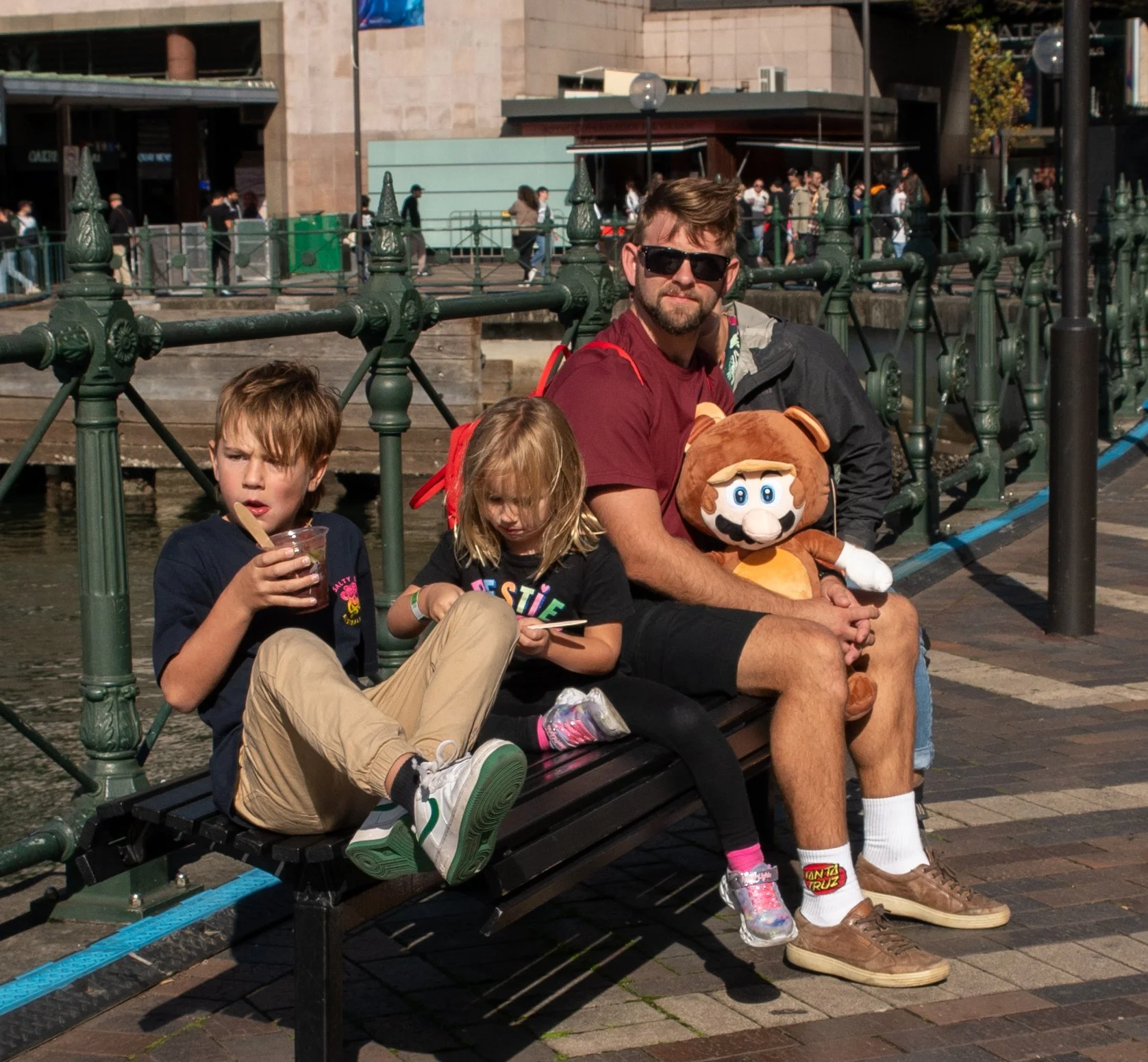 A man and two children sitting on a park bench by a waterfront in an urban area, with the man holding a Mario plush toy and wearing sunglasses.