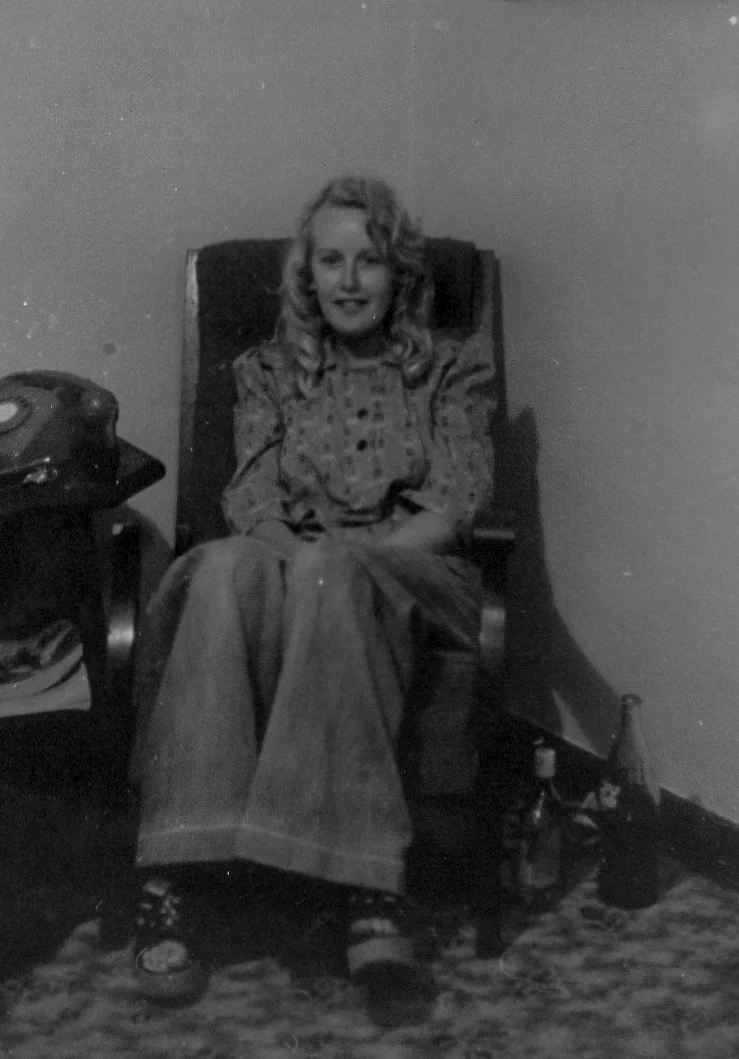 A black-and-white photo of a woman sitting in a wooden armchair, smiling. She has curly hair and is wearing a patterned blouse and wide trousers. There is a rotary phone on a small table beside her, and a glass bottle on the floor nearby.