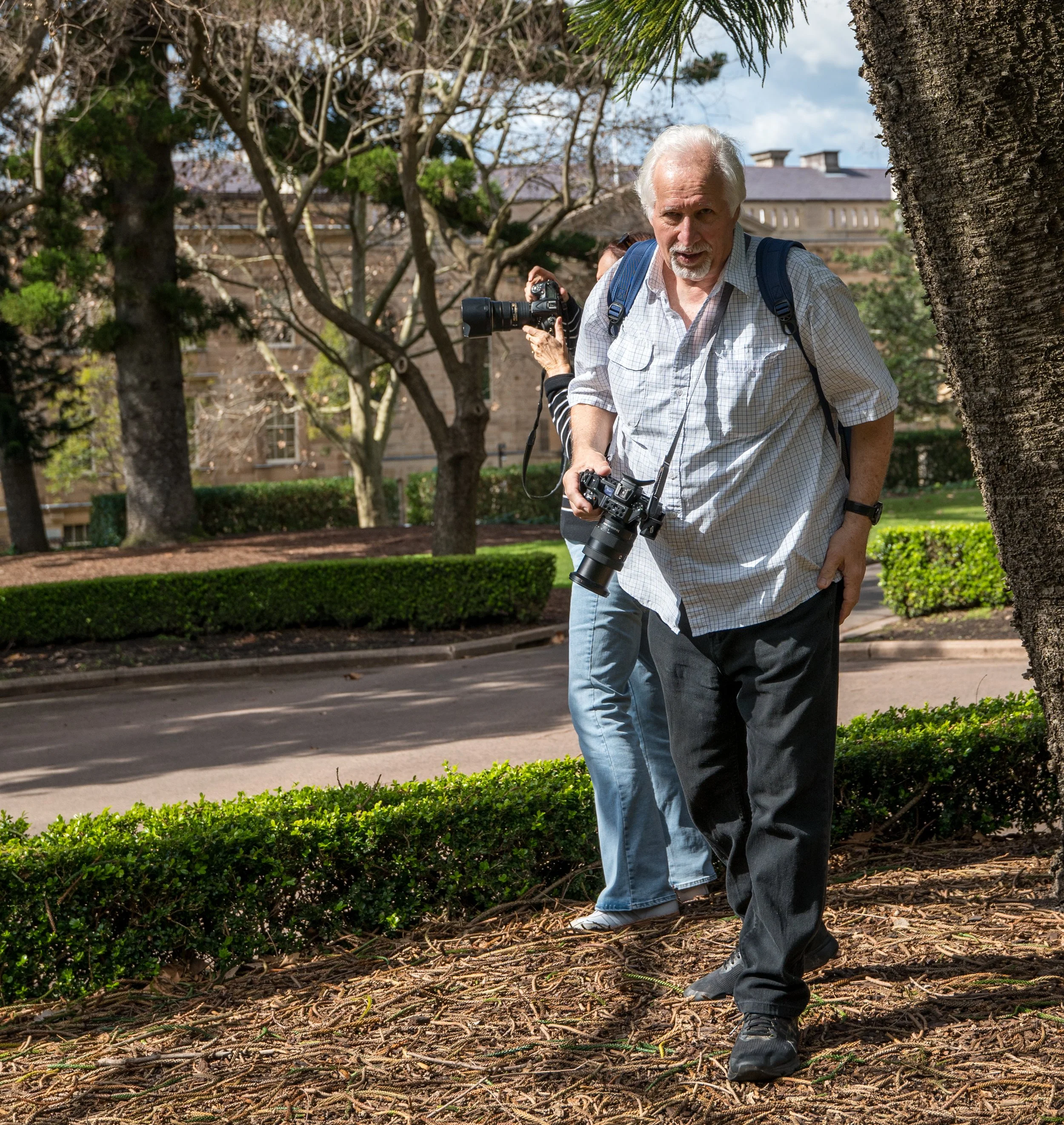 Two men walking in a park, carrying cameras, with trees and a building in the background.