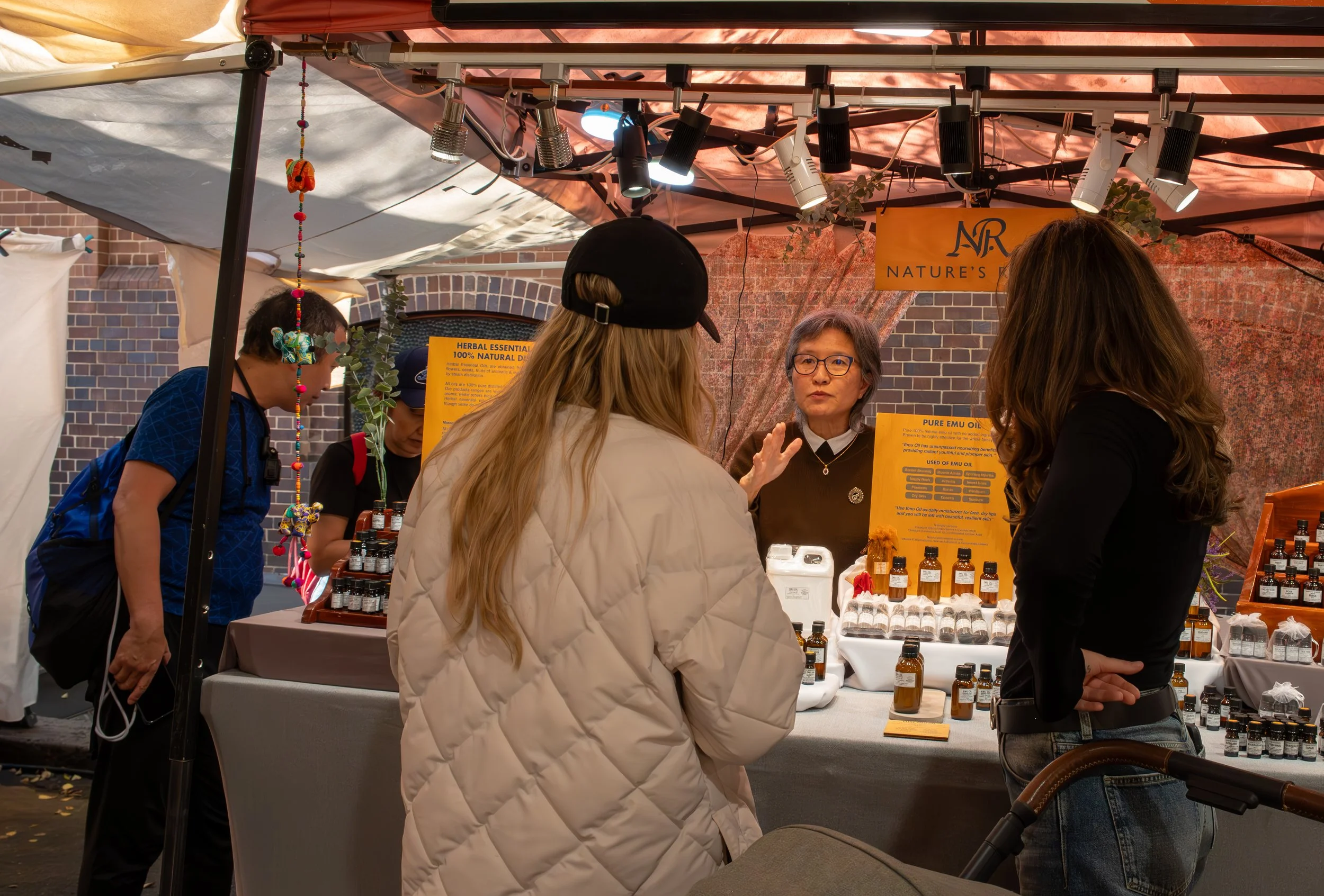 A vendor at a market stall explaining products to three women. The stall displays bottles of essential oils with a yellow sign and a colorful string of decorations hanging from the tent. The background features a brick wall and the stall has a banner with the words "Nature's." One woman has long blonde hair and wears a black cap, another with long brown hair wears a black top, and the third woman has short gray hair and glasses.