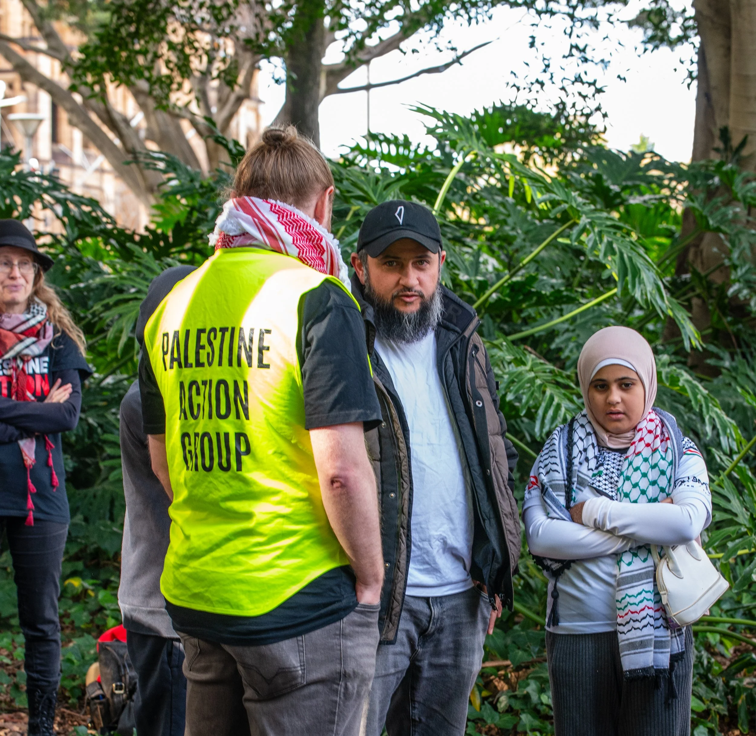 A group of people gathered outdoors among lush green trees, including a man with a beard in a black jacket, a woman in a hijab, and a person wearing a yellow vest with 'Palestine Action Group' written on it, speaking to the others.