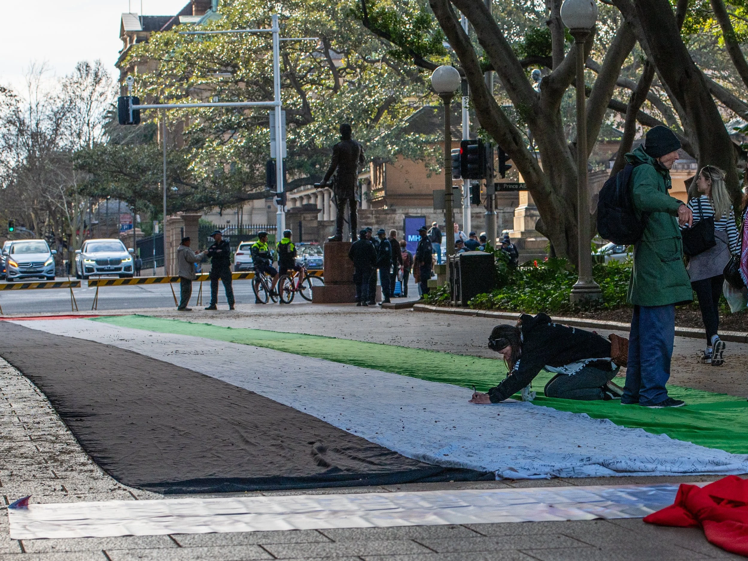 People painting a large flag on the sidewalk with green, white, and black colors, while others watch and take photos. Police and cyclists are visible in the background.