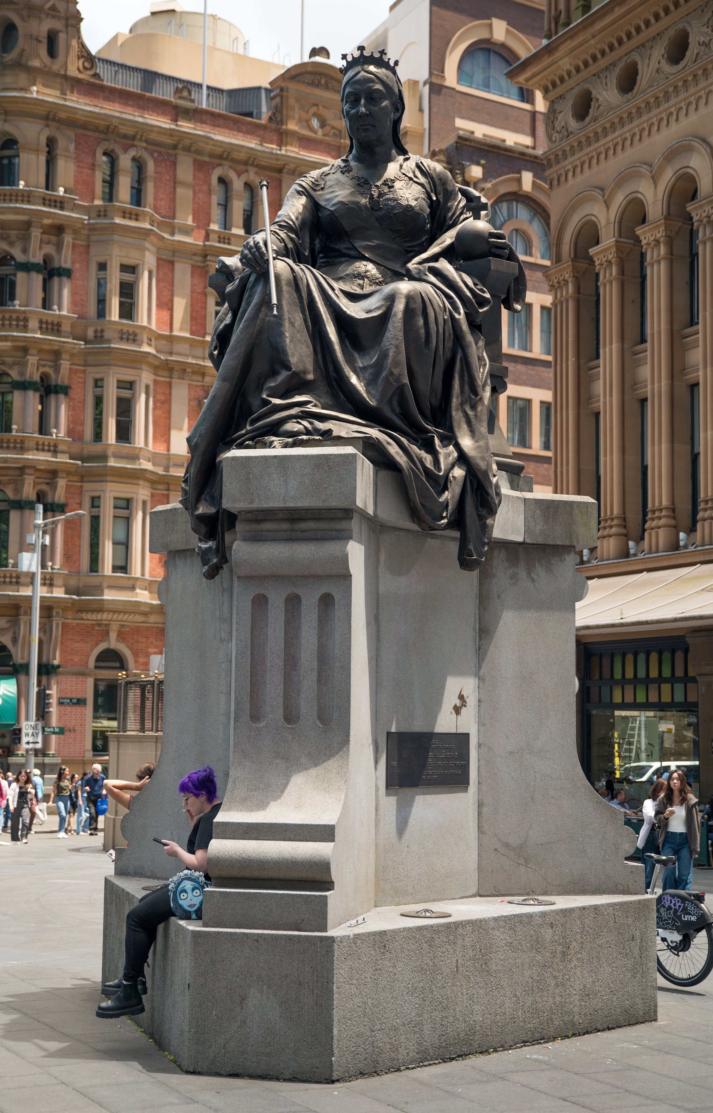 A statue of a woman seated on a throne-like chair in an urban setting, surrounded by historic buildings and pedestrians, with a person sitting at the base looking at their phone.