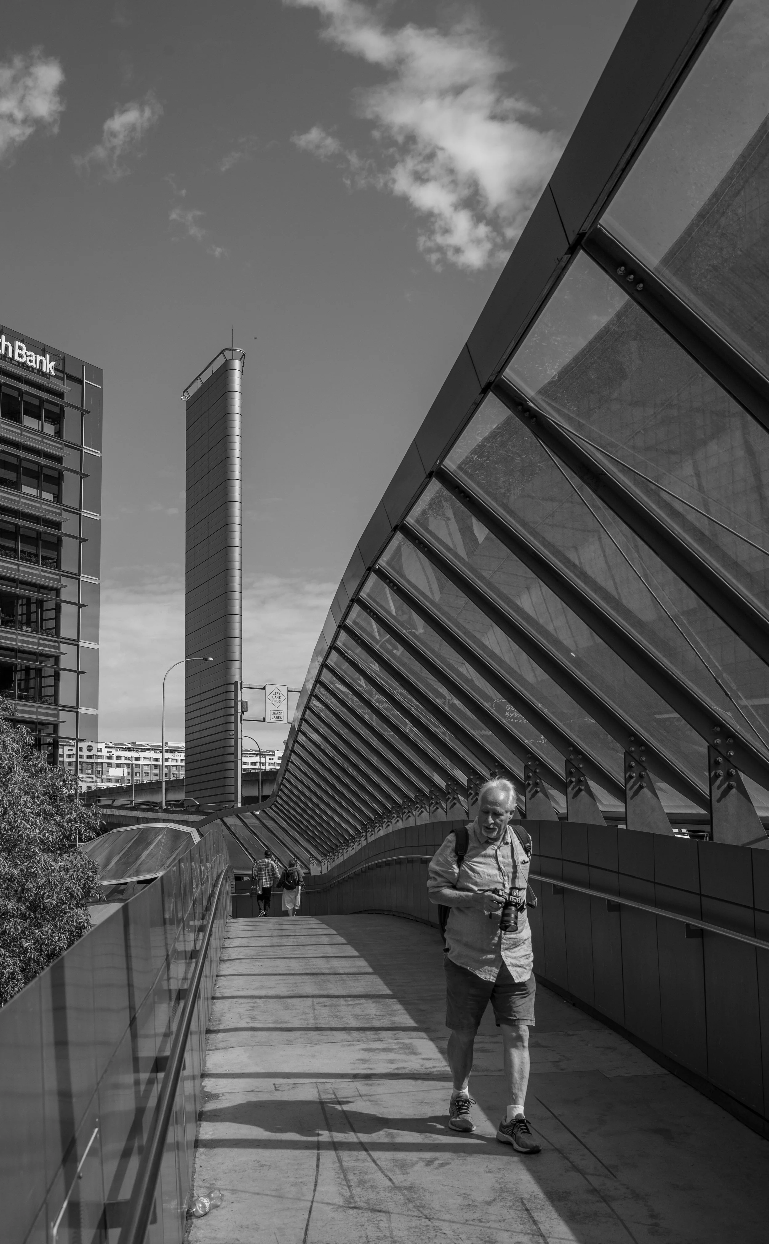 A black and white photo of an urban scene with modern buildings, including a tall cylindrical skyscraper, and a man walking with a camera on a pedestrian bridge with glass and metal architecture.